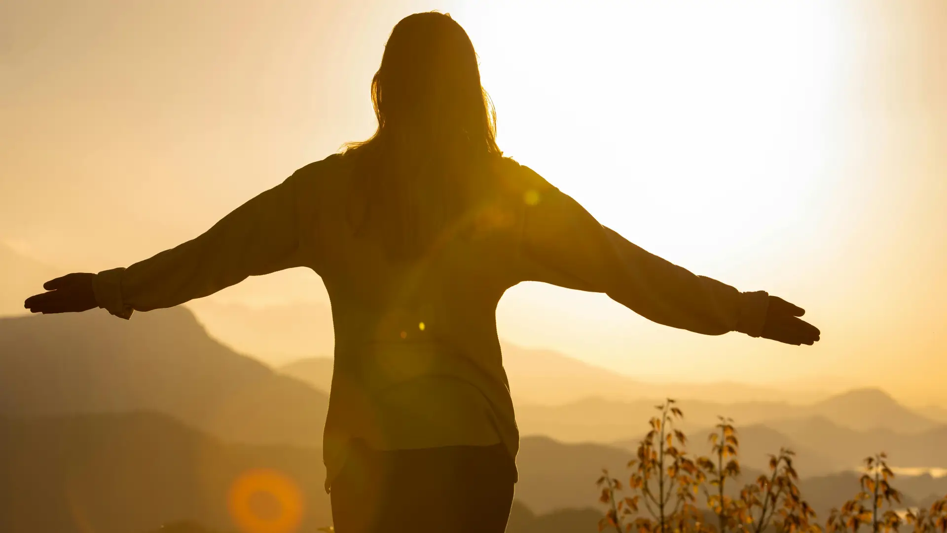 Woman's silhouette with outstretched arms in front of distant mountain view at golden sunrise.