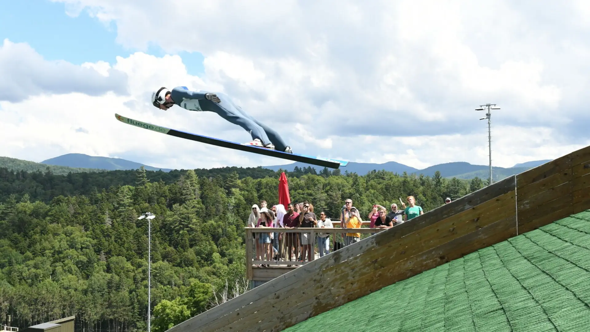 Ski jumper takes off of turf surface in summer while observation deck full of people looks on