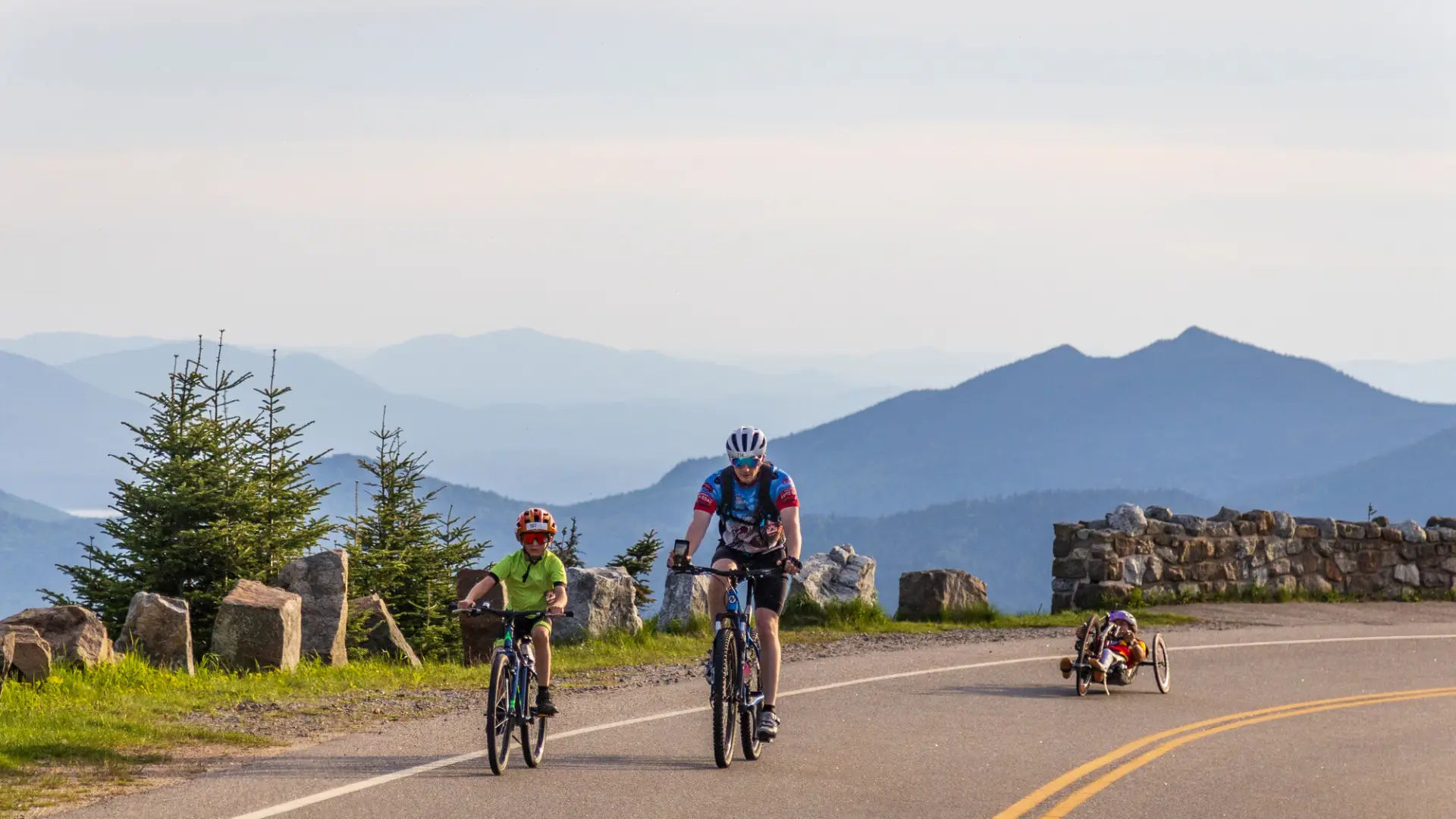 A cyclist and a child on a bicycle ride along a road with scenic views, followed by another person in a recumbent handbike.