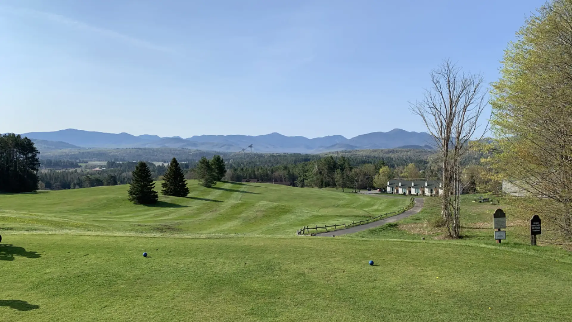 Tee and fairway at Lake Placid Club golf course, with Olympic ski jumps and Adirondack peaks in the distance.