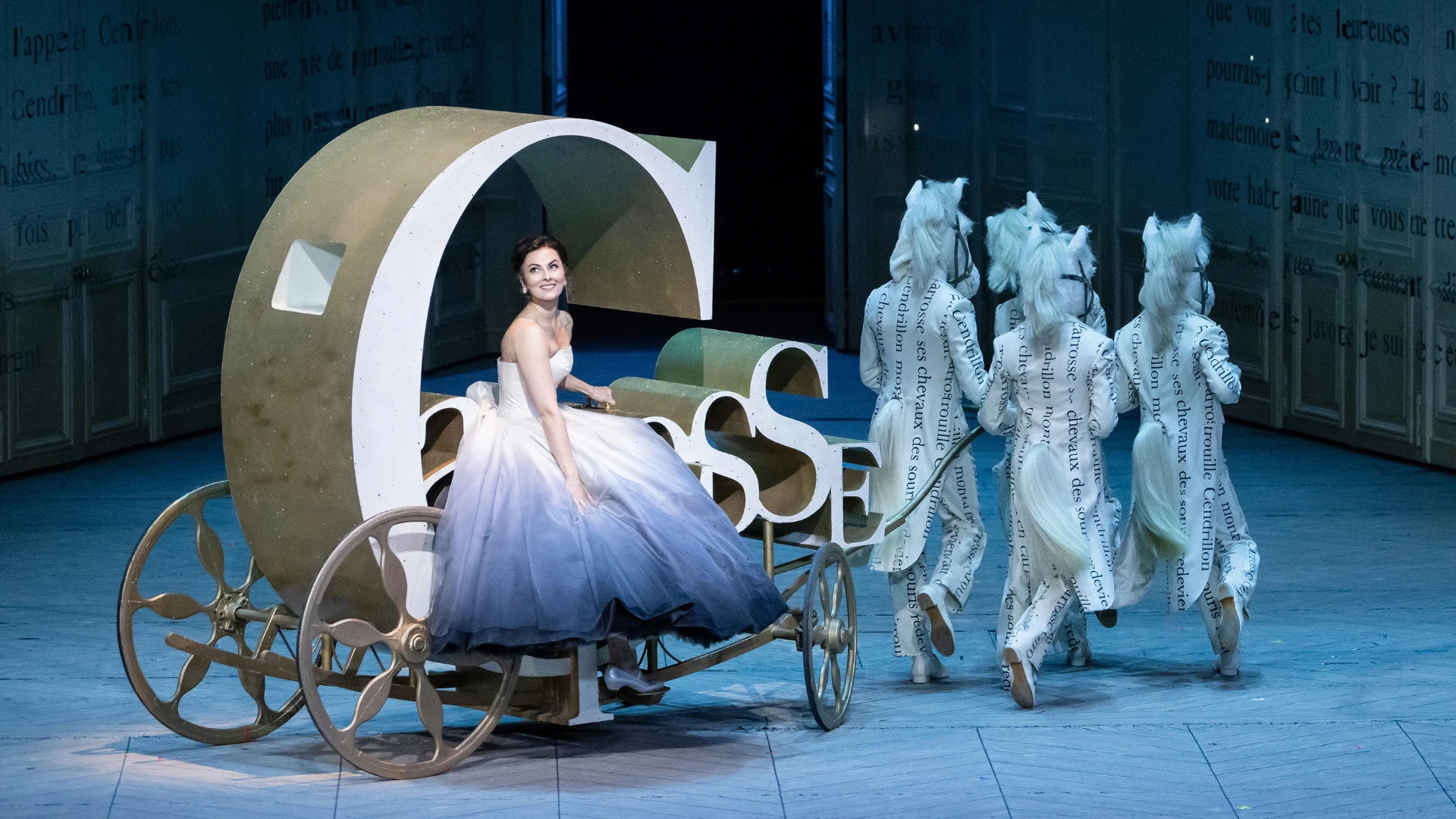 Woman in evening dress sitting in carriage on stage pulled by four people dressed in suits and horse heads