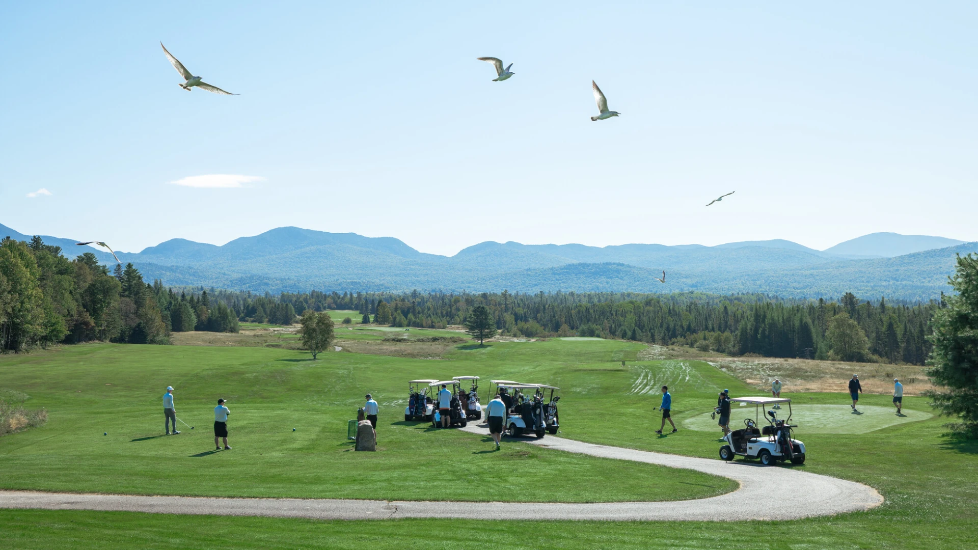 Golfers and golf carts on green course with mountains in distant background.
