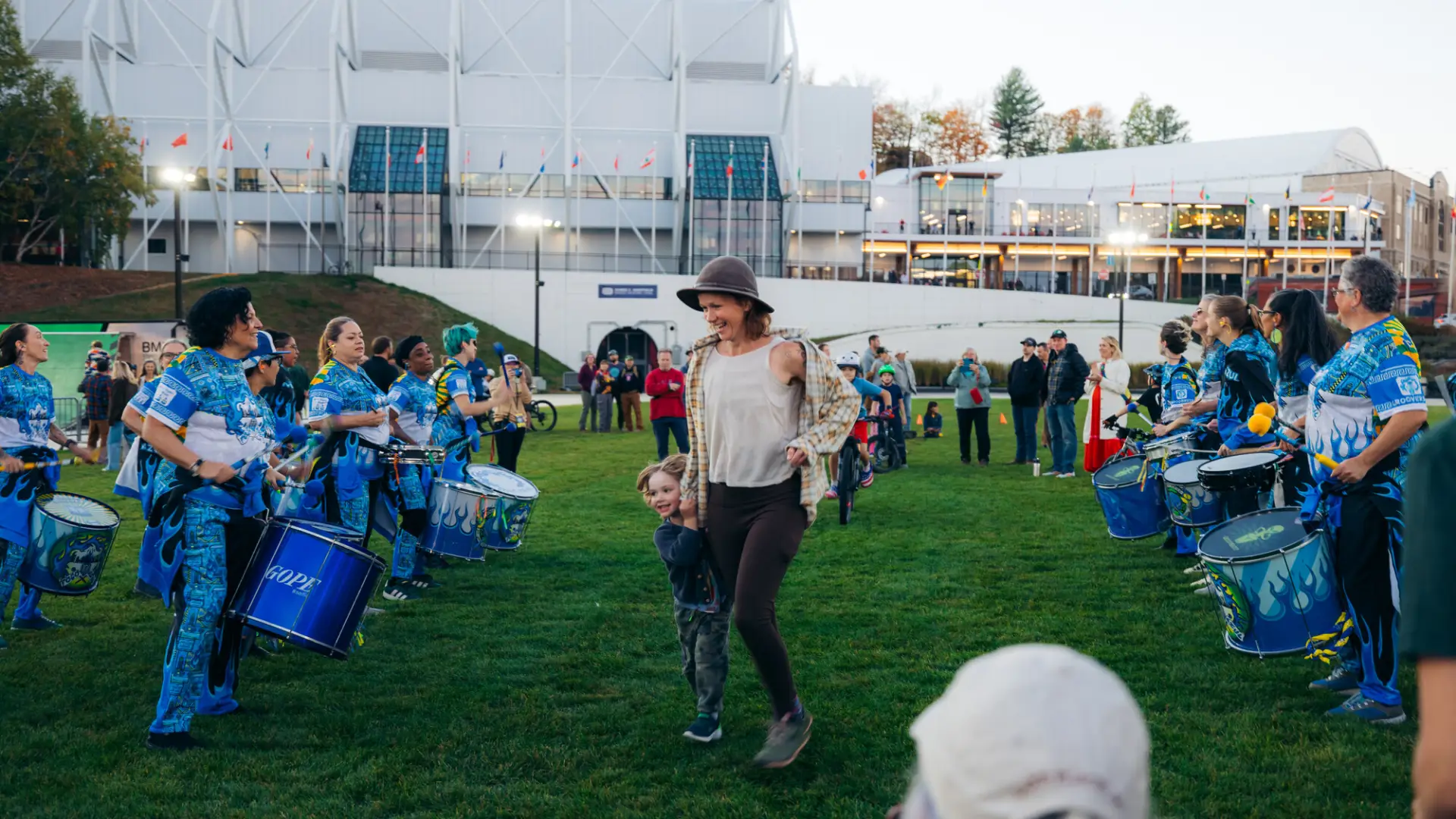 Woman holds child's hand as they run through the middle aisle of a drum line on the Olympic Oval in Lake Placid