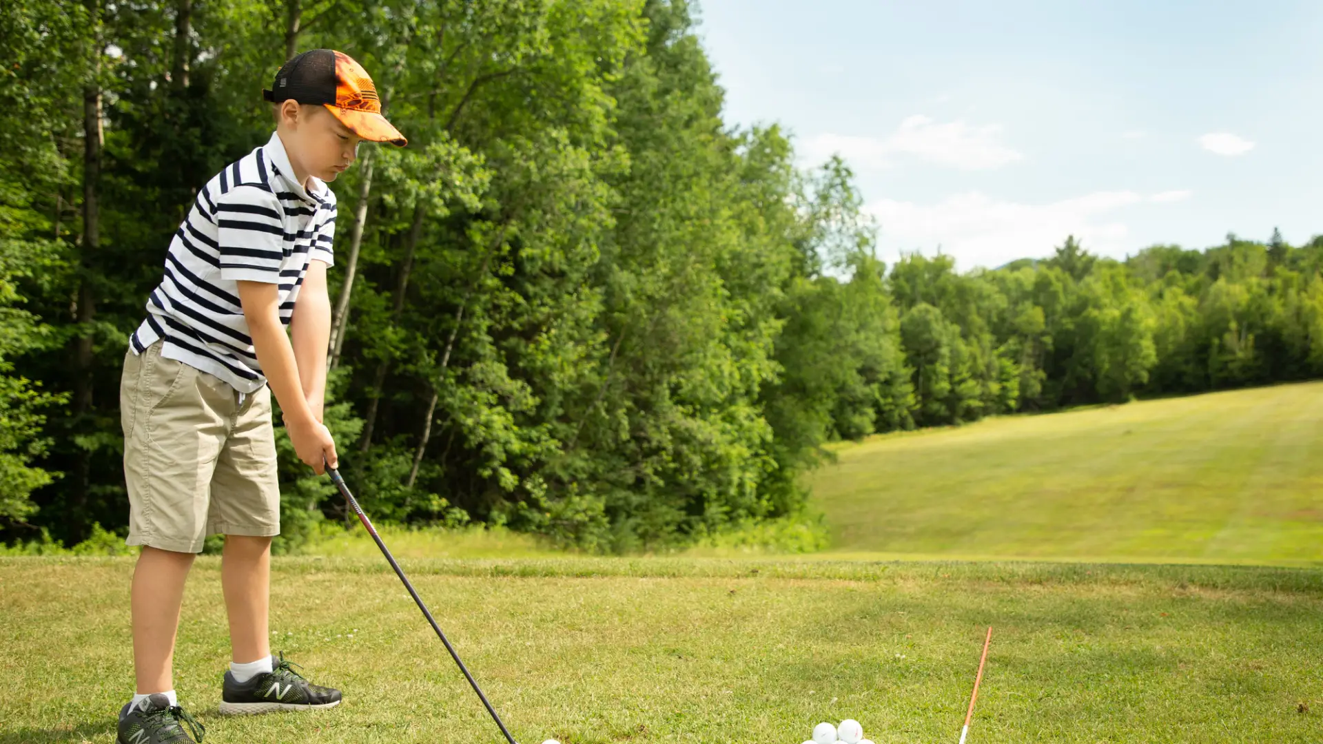 A young boy prepares to tee off on a golf green, with bright green trees along the fairway.