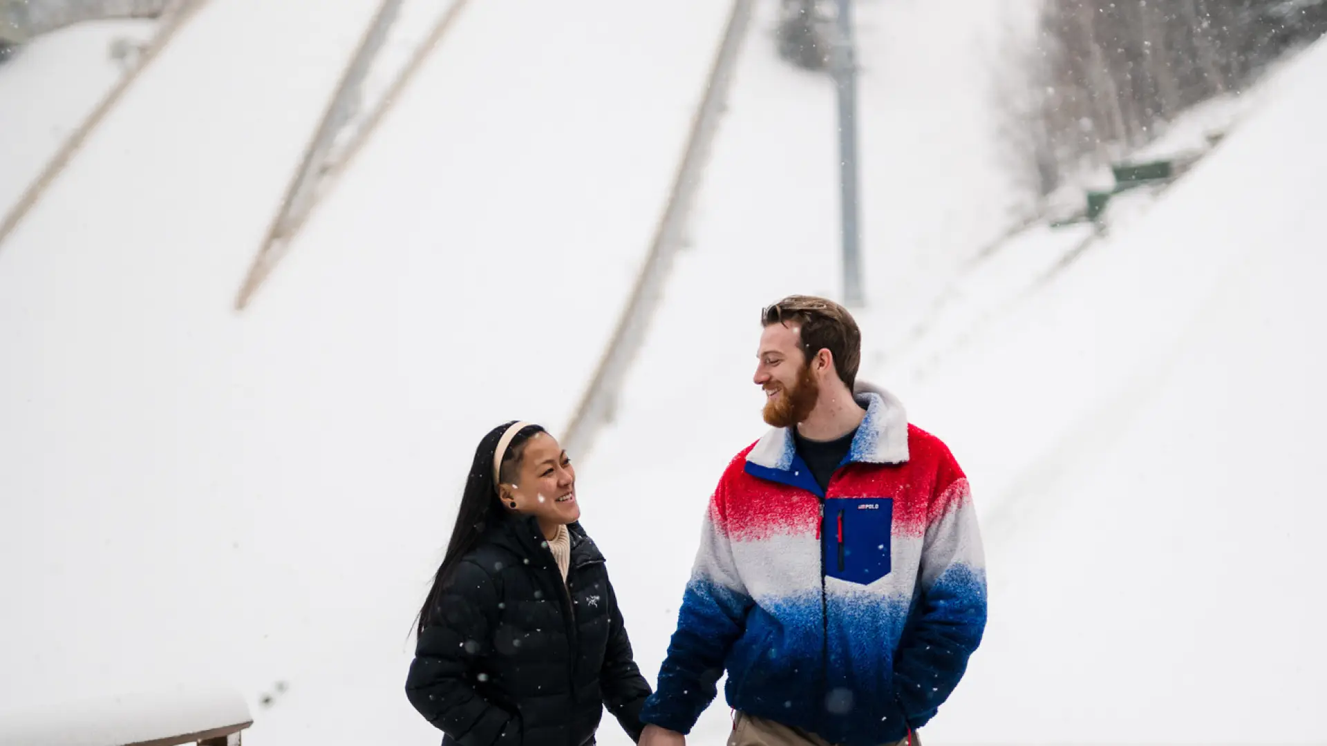A couple holds hands and smiles at each other in front of snow-covered ski jumps.