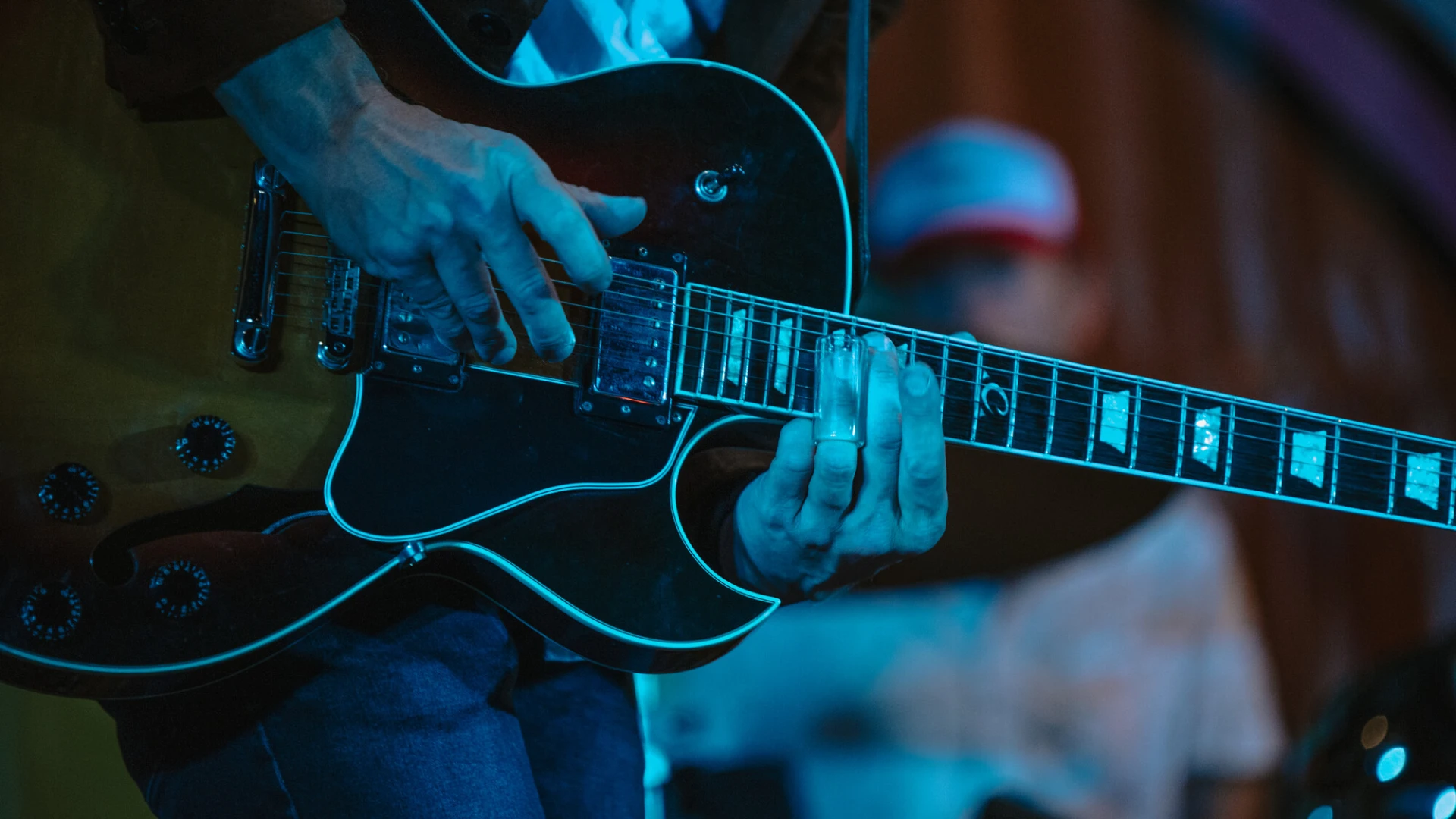 Close-up of person playing electric guitar on dark stage with drummer in background