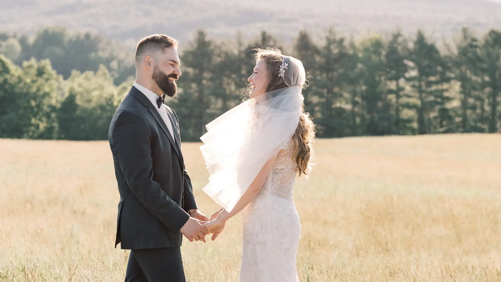 Bride and groom smile at each other in a wheaty field with veil blowing in the breeze