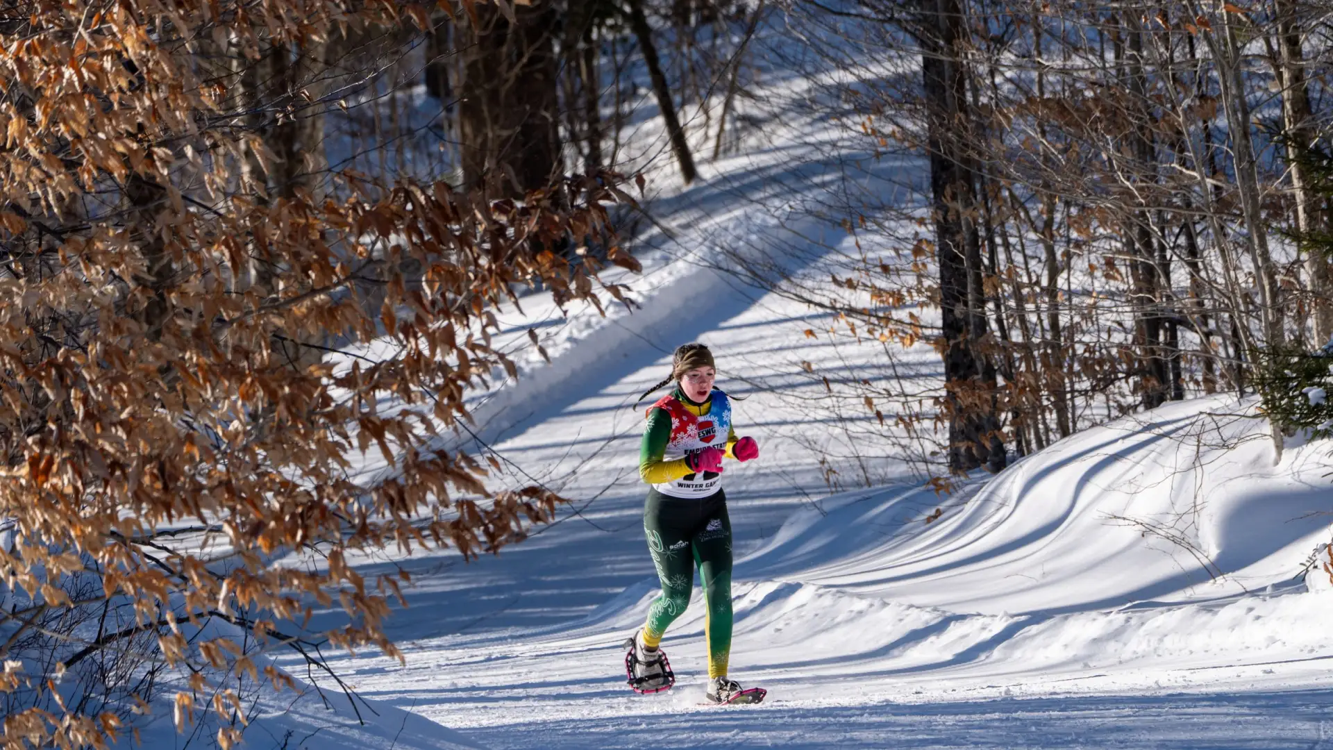 A young woman snowshoeing on a snowy trail, wearing vibrant athletic gear, under a clear blue sky with trees in the background.