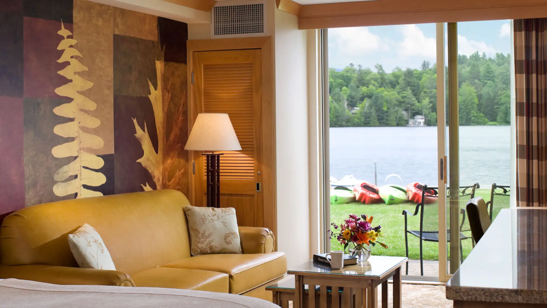 A hotel room with sliding doors opening onto a lakefront lawn with kayaks.