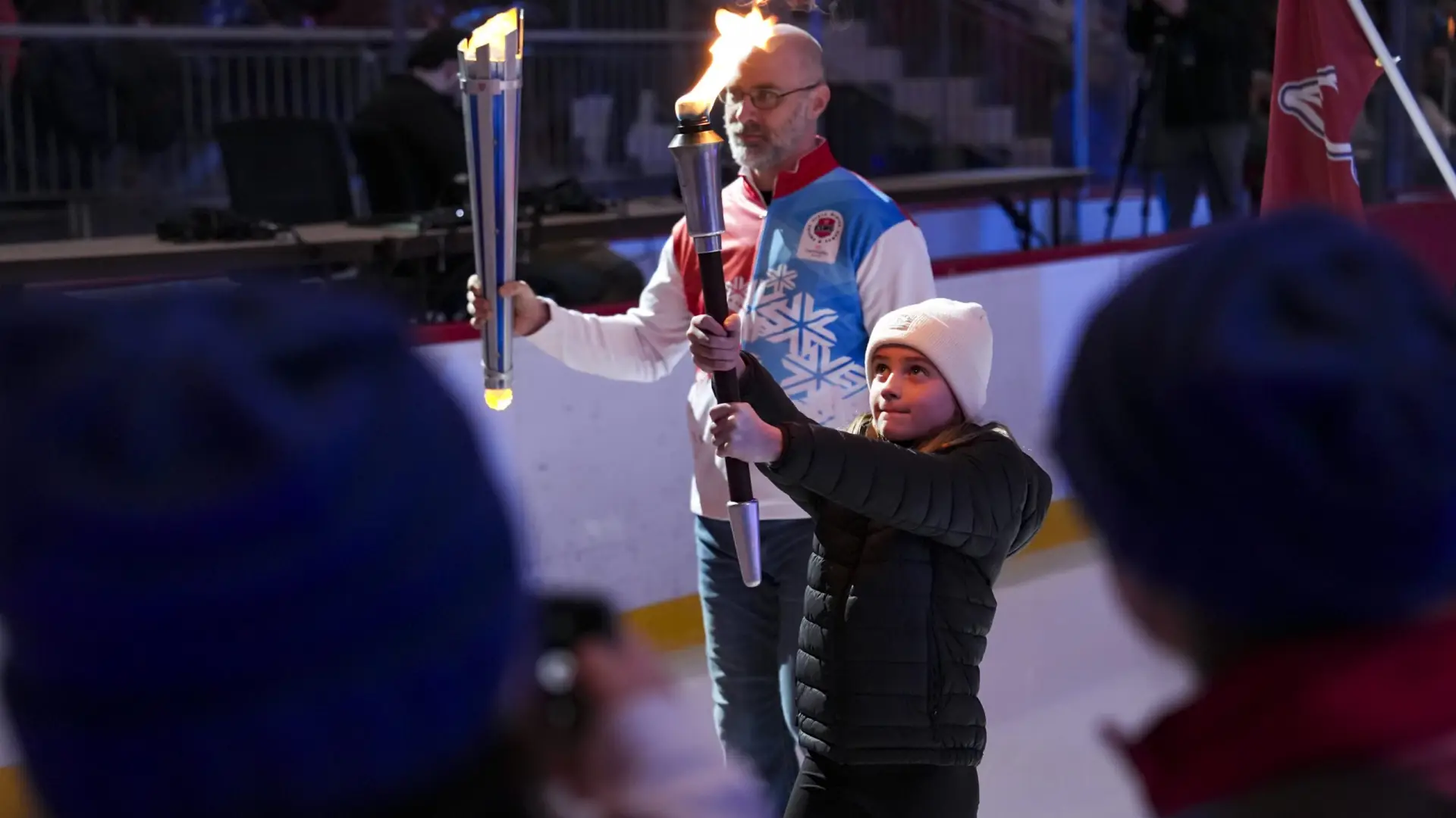 A young girl in a white beanie holds a flaming torch, assisted by an older man in a blue and red jacket. They stand indoors on an ice rink, surrounded by onlookers. The scene feels ceremonial and celebratory.
