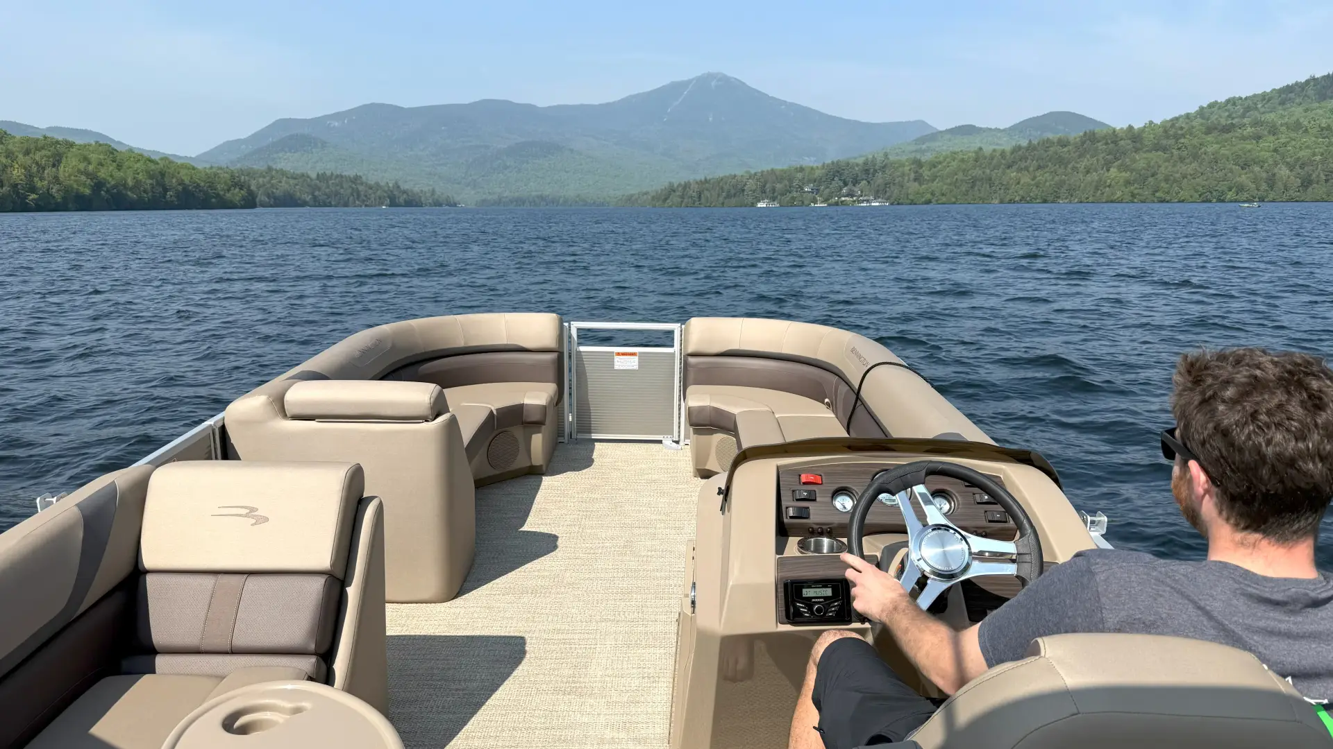Man drives a pontoon boat on a lake towards a mountain in the distance