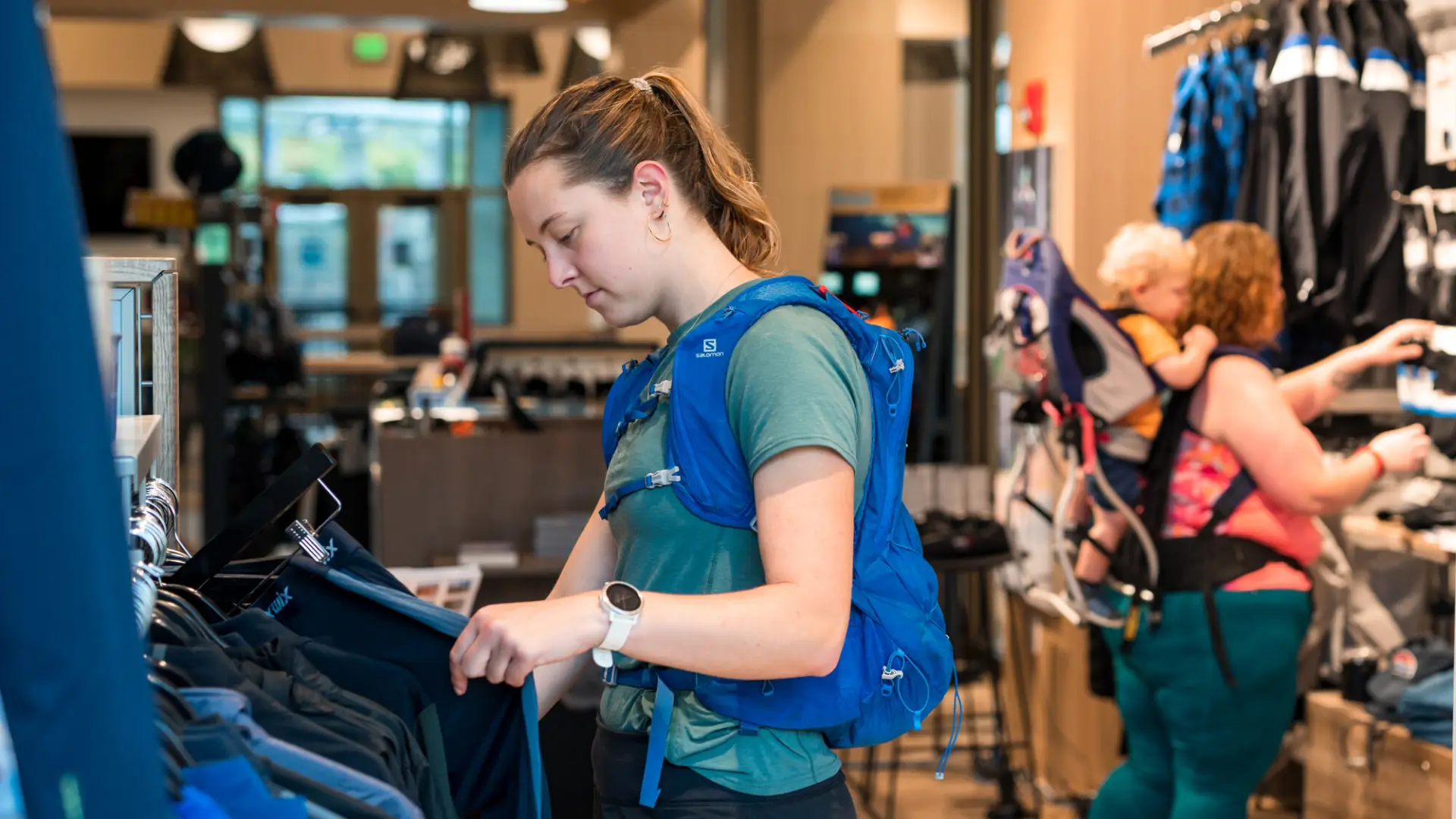 A young woman wearing a hiking water pack browses a rack in an outdoor gear outfitters.