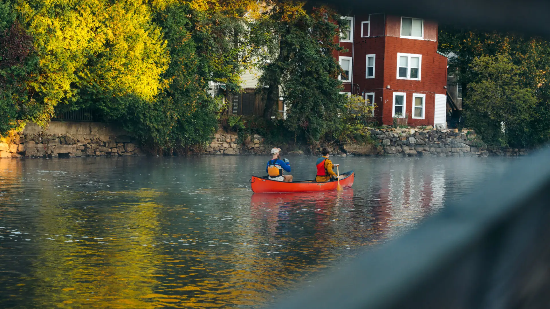 Man and woman paddle a red canoe down a river with a red house and trees on the bank