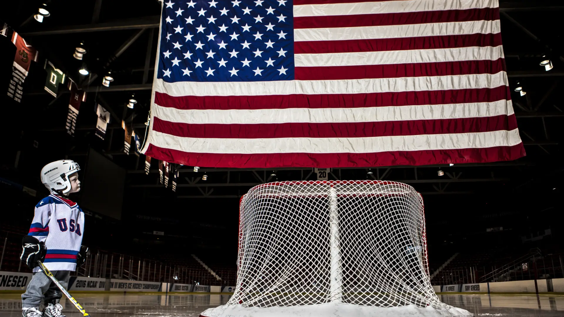 Young boy in hockey gear stands next to hockey goal faced away with large American flag flying above it in hockey rink on ice