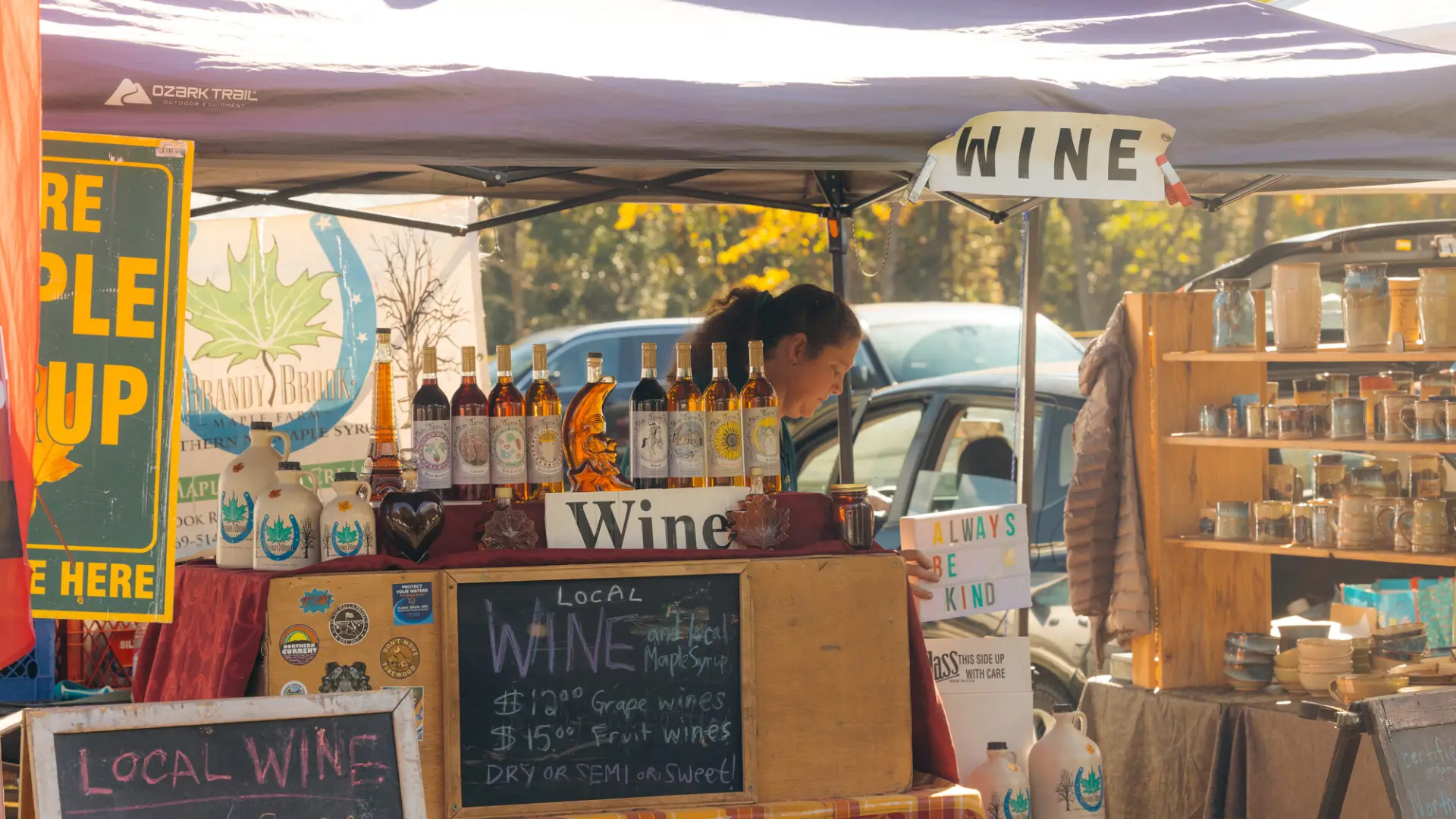 Woman holding signs that says "always be kind" stands behind wine display decorated with chalkboard signs under purple tent at farmers market