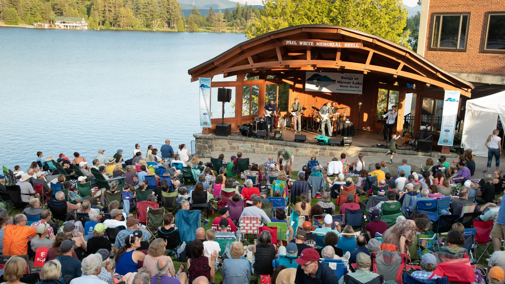 A crowd gathers near the band shell in the grass under a summer sky at Mid's Park in Lake Placid