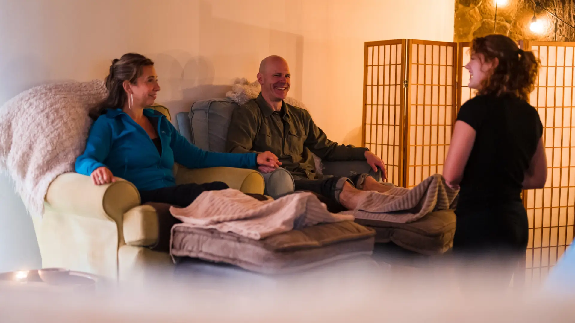 A man and woman sit next to each other on plush armchairs, with their feet up for a spa treatment from a woman with curly hair.