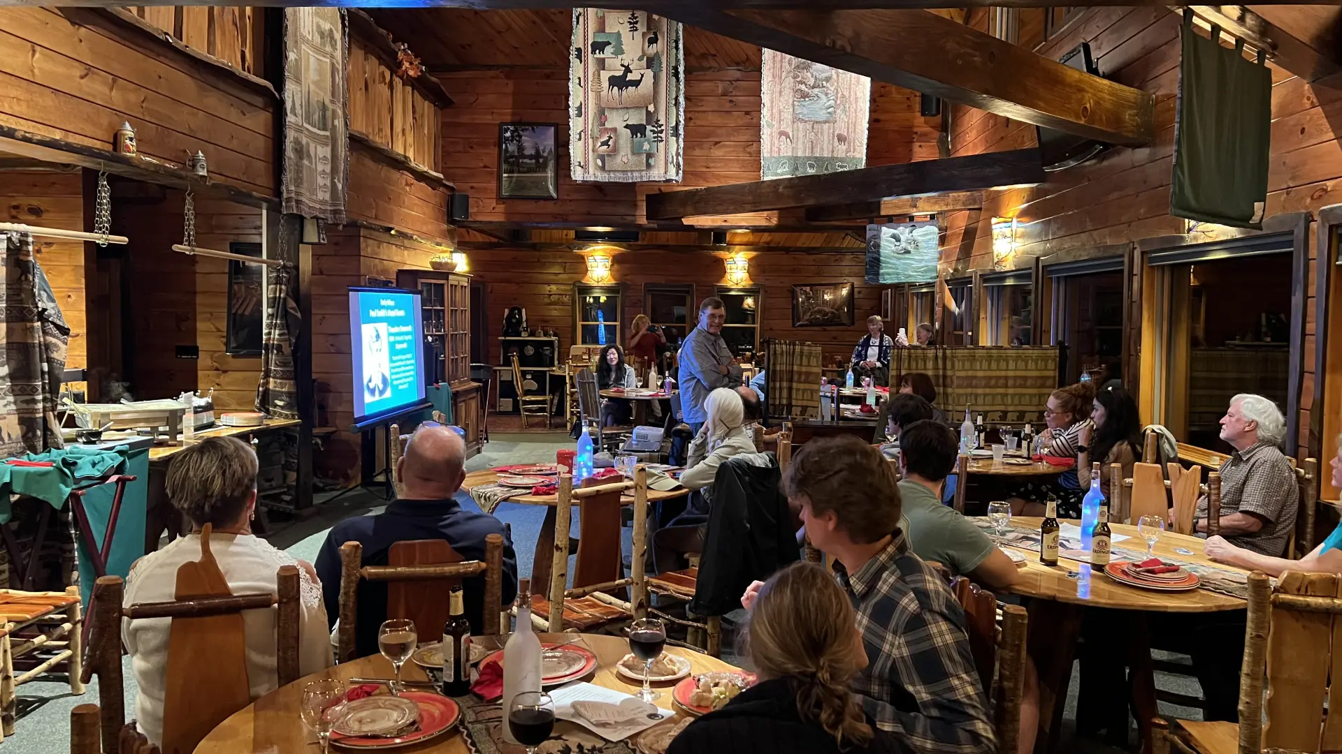 A group of people seated at round tables in a rustic wooden lodge listen to a presentation shown on a screen.