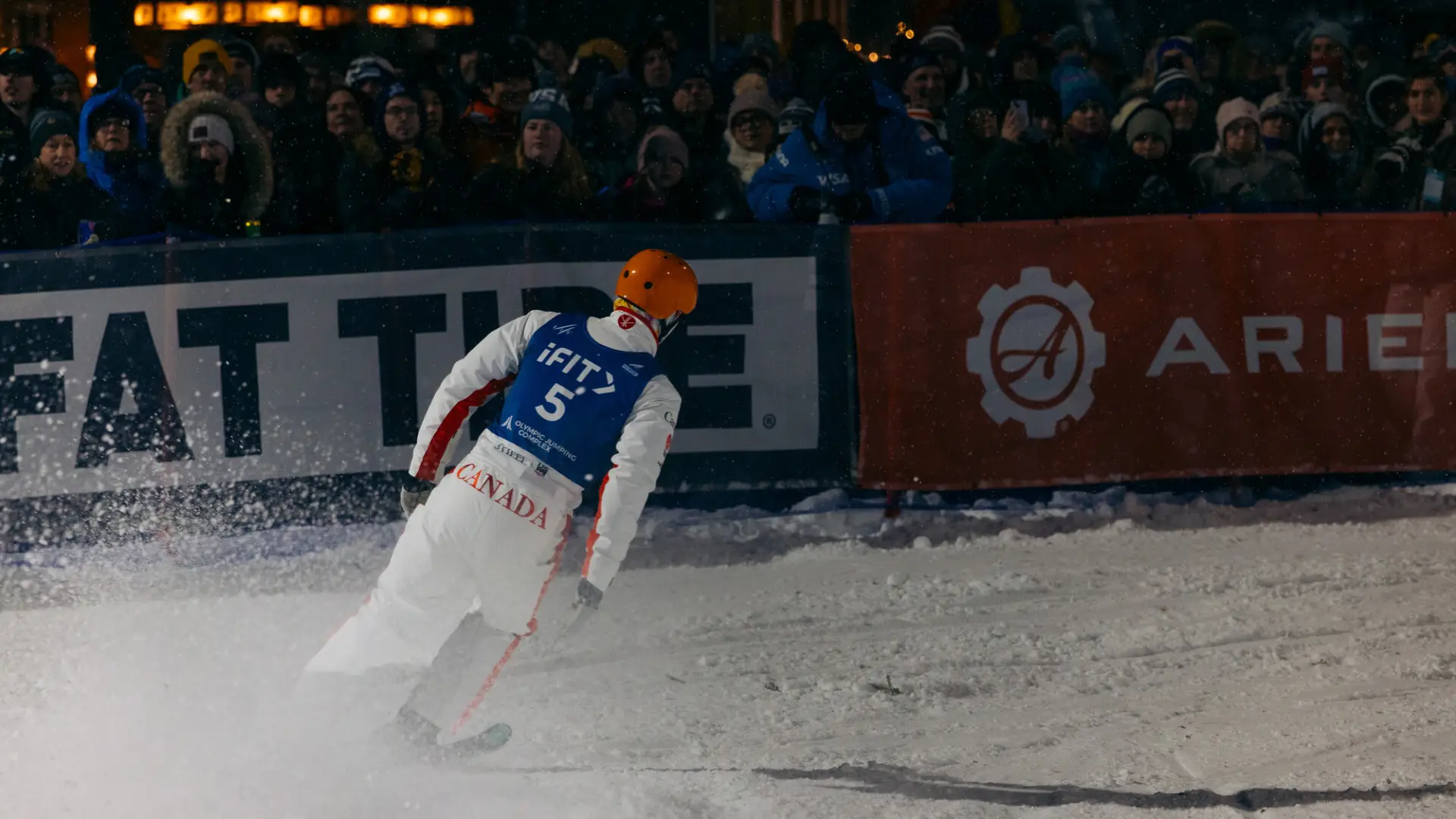 Freestyle aerialist skier skis into finish while a crowd looks on over a barrier at nighttime event
