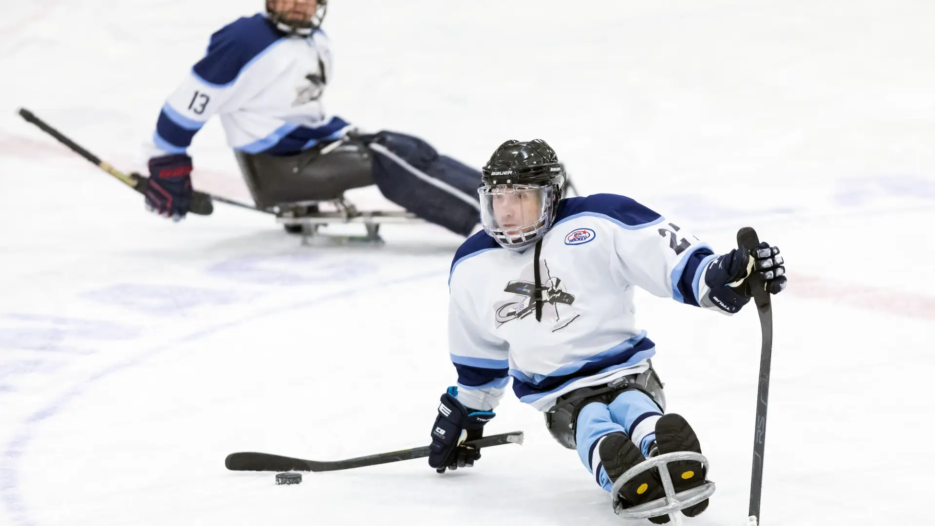 A close-up action shot of two para ice hockey (sled hockey) players on an indoor rink during the Empire State Winter Games.