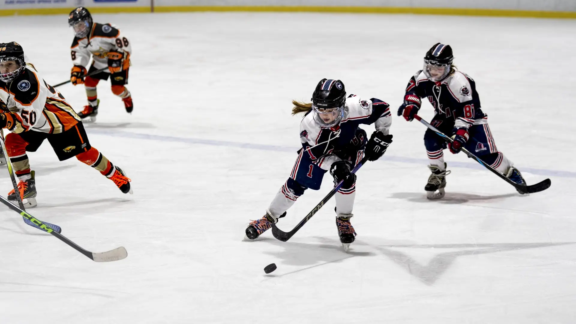 Exciting U10 girls' ice hockey match: a focused player in a white and navy uniform skates with the puck, pursued by teammates and opponents.