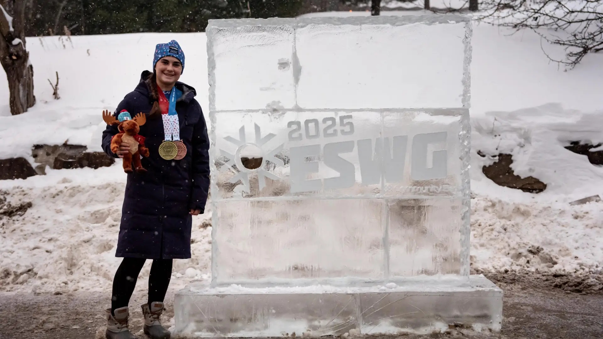 A smiling athlete stands proudly by a large ice sculpture for the 2025 ESWG, holding medals and a plush moose toy.