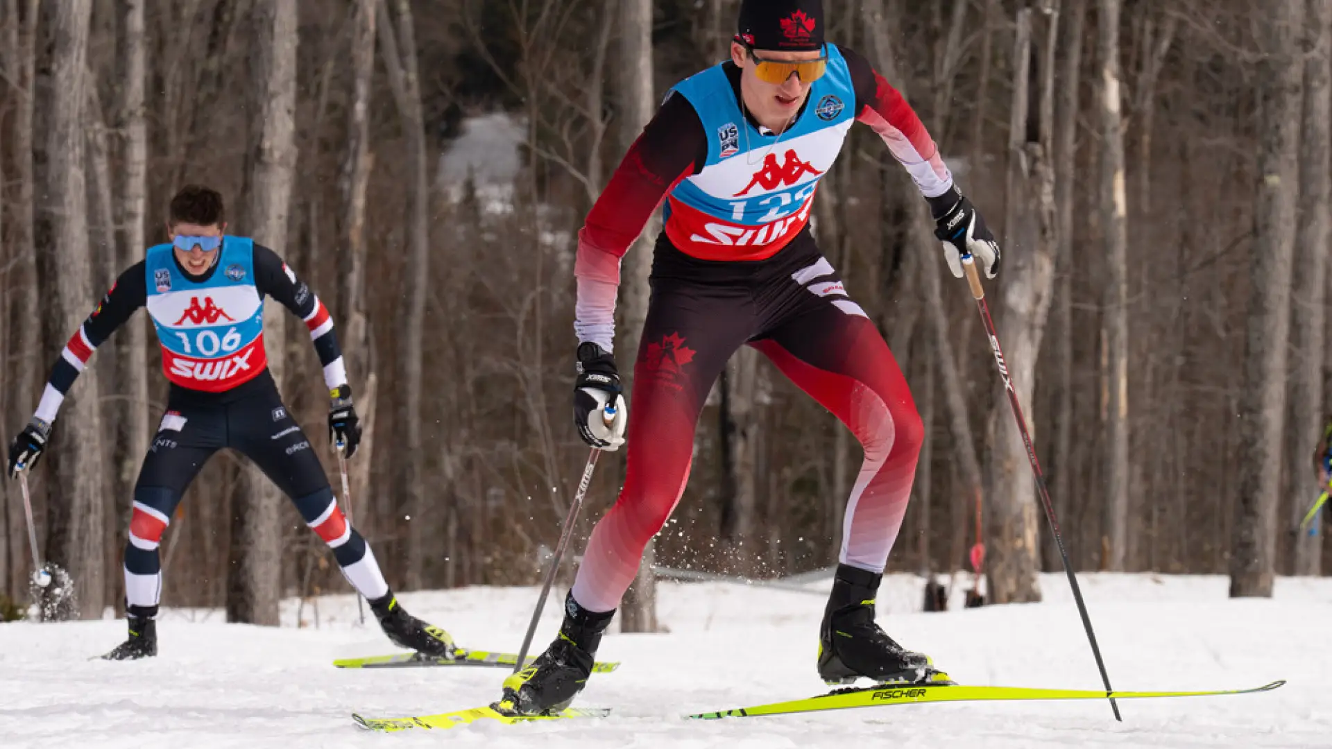 The image shows two cross-country skiers racing on a snowy trail. 