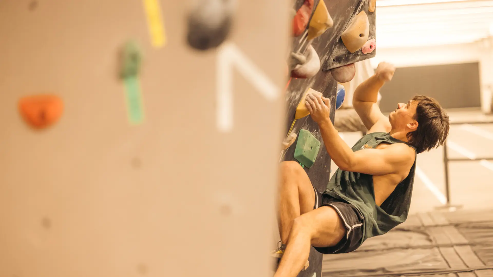 Bouldering wall with colorful tape next to holds in foreground, with a climber seen on the wall around a corner