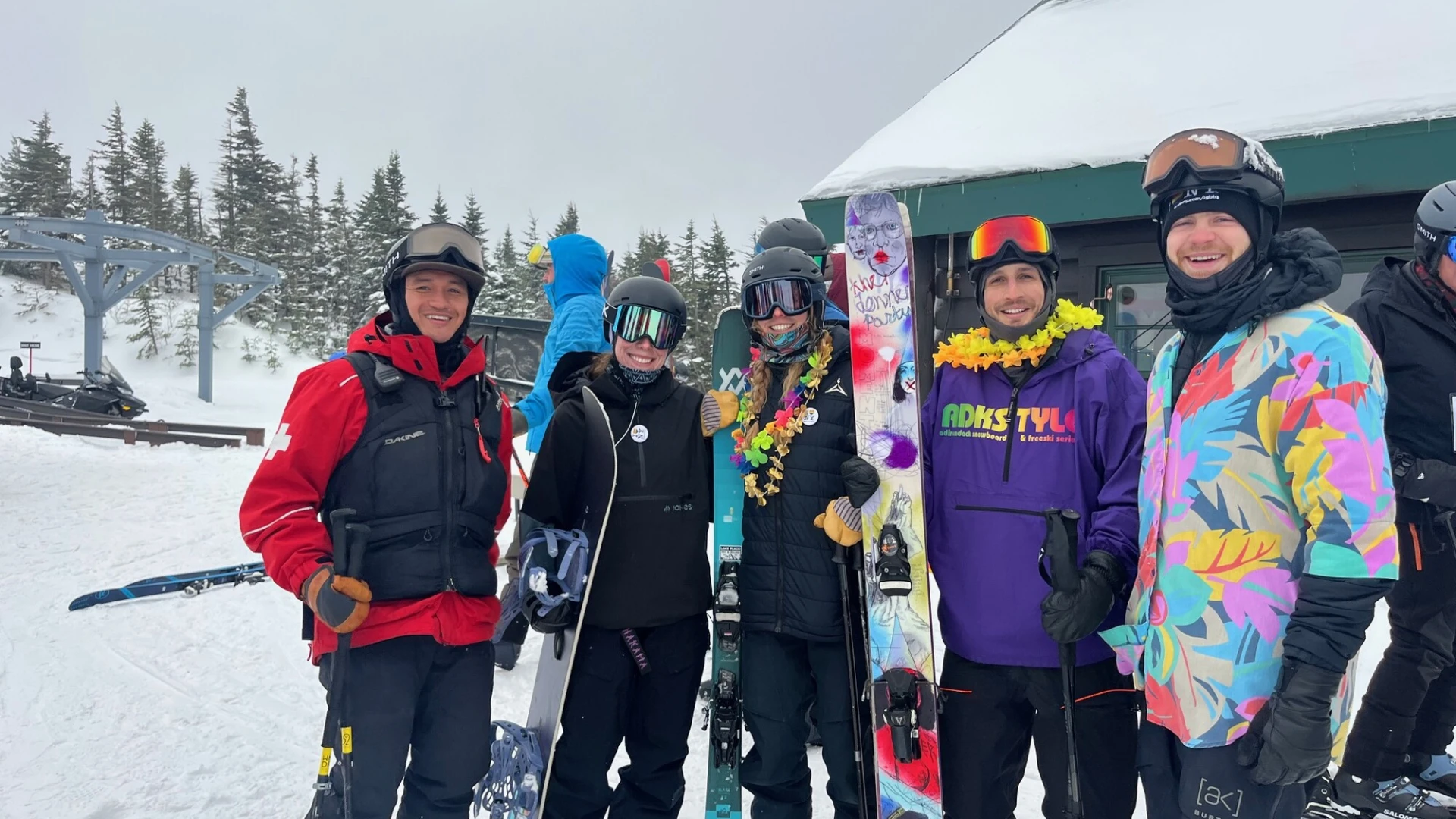 A group of skiers and snowboarders, some wearing colorful leis, pose together near a mountain building.