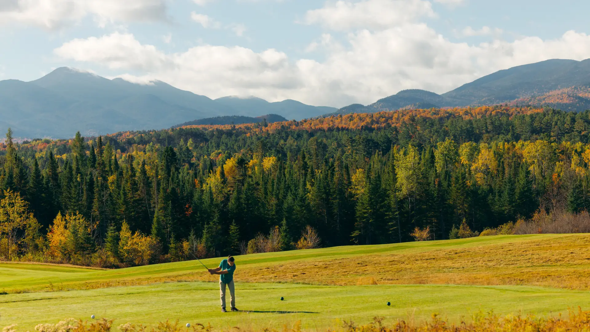 A golfer tees off on a green course, with soaring fall foliage-covered mountains in the background.