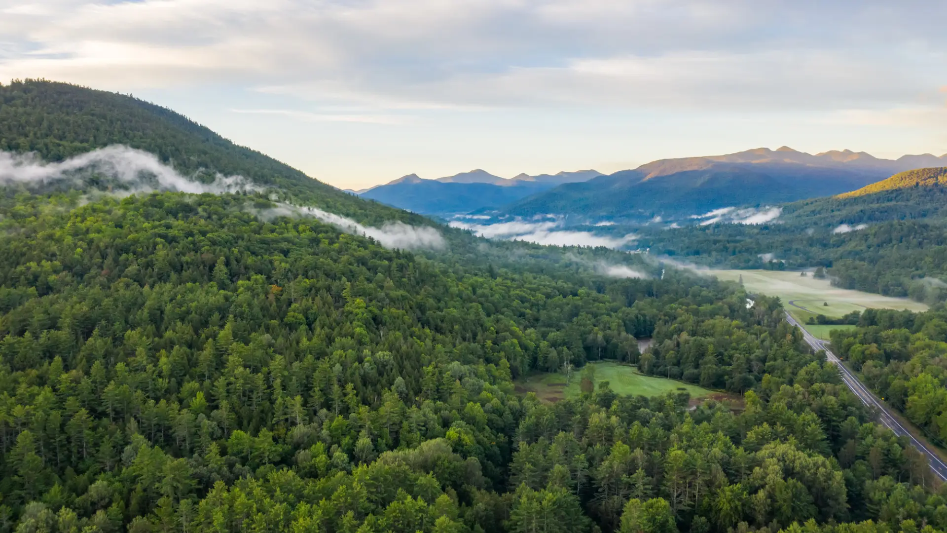 An aerial view of a forested mountain with a road and more mountains in the distance. 