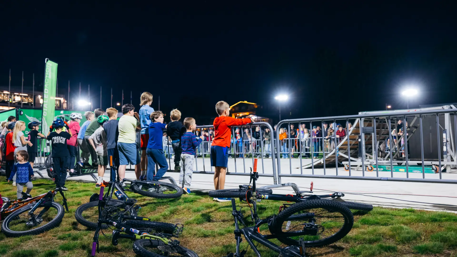Bikes litter the lawn and children stand against event fencing watching BMX stunt show on Olympic Oval
