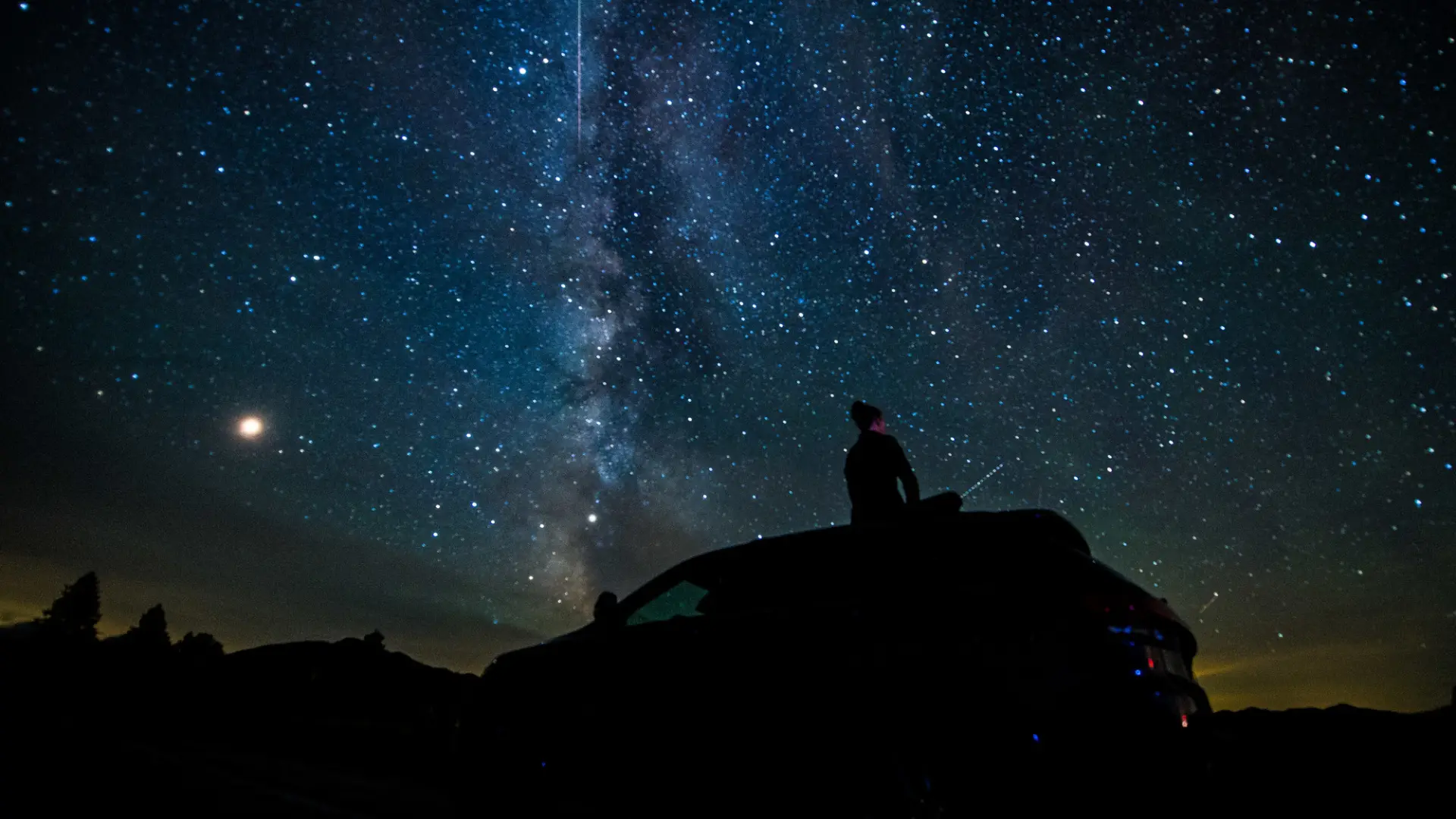 Person sits on the top of a vehicle viewing the stars under the Adiroddack night sky.
