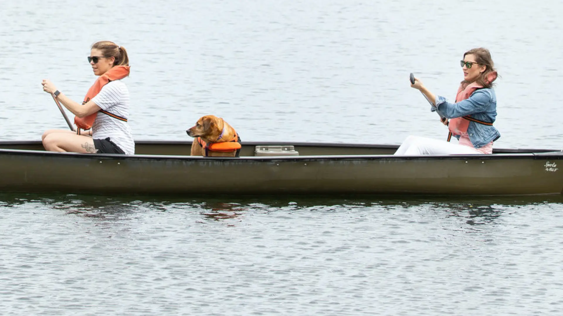 Two women and a dog in a brown canoe.