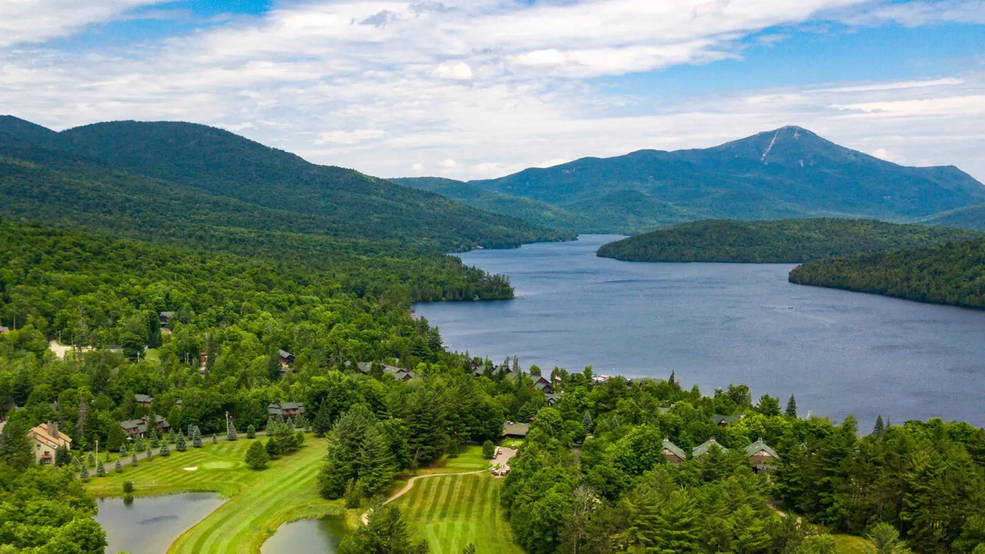 An aerial view of a golf course on a lakeshore with mountains in the background.