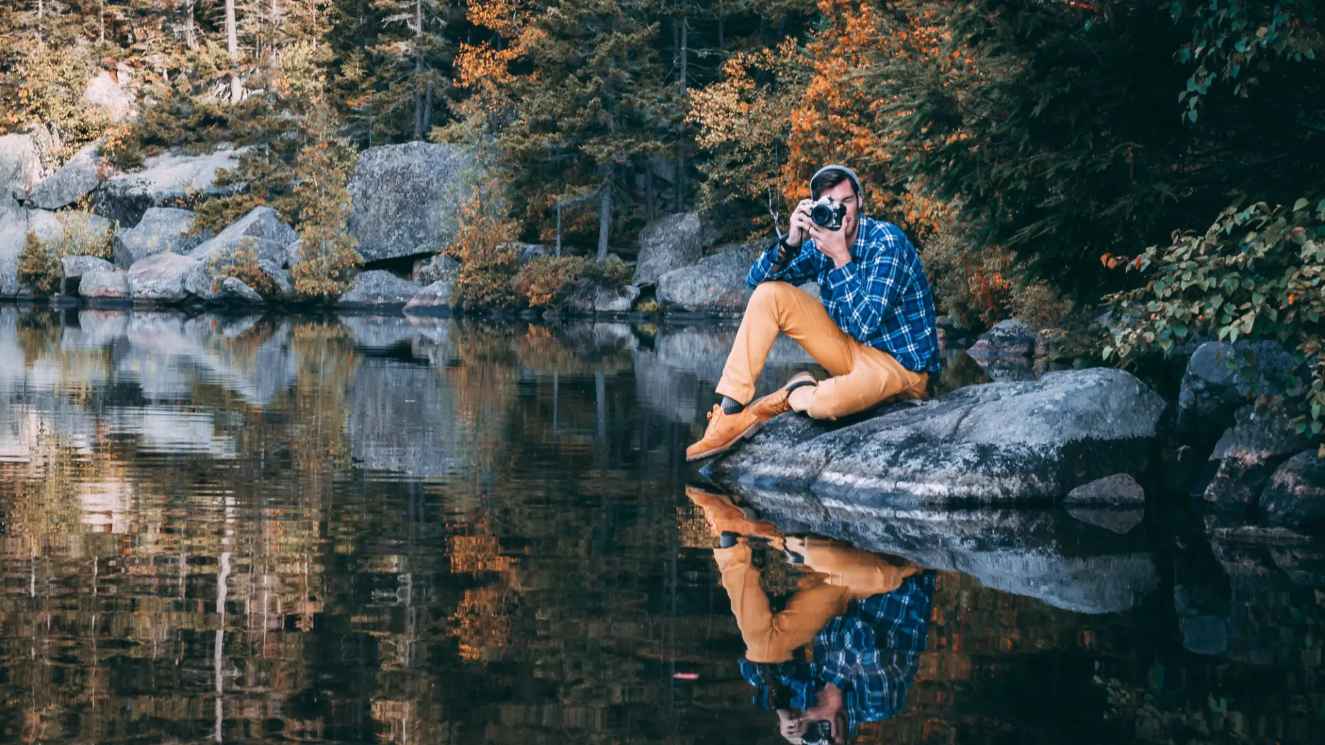 A person siting on a rock by the water holding a camera with fall foliage behind them.