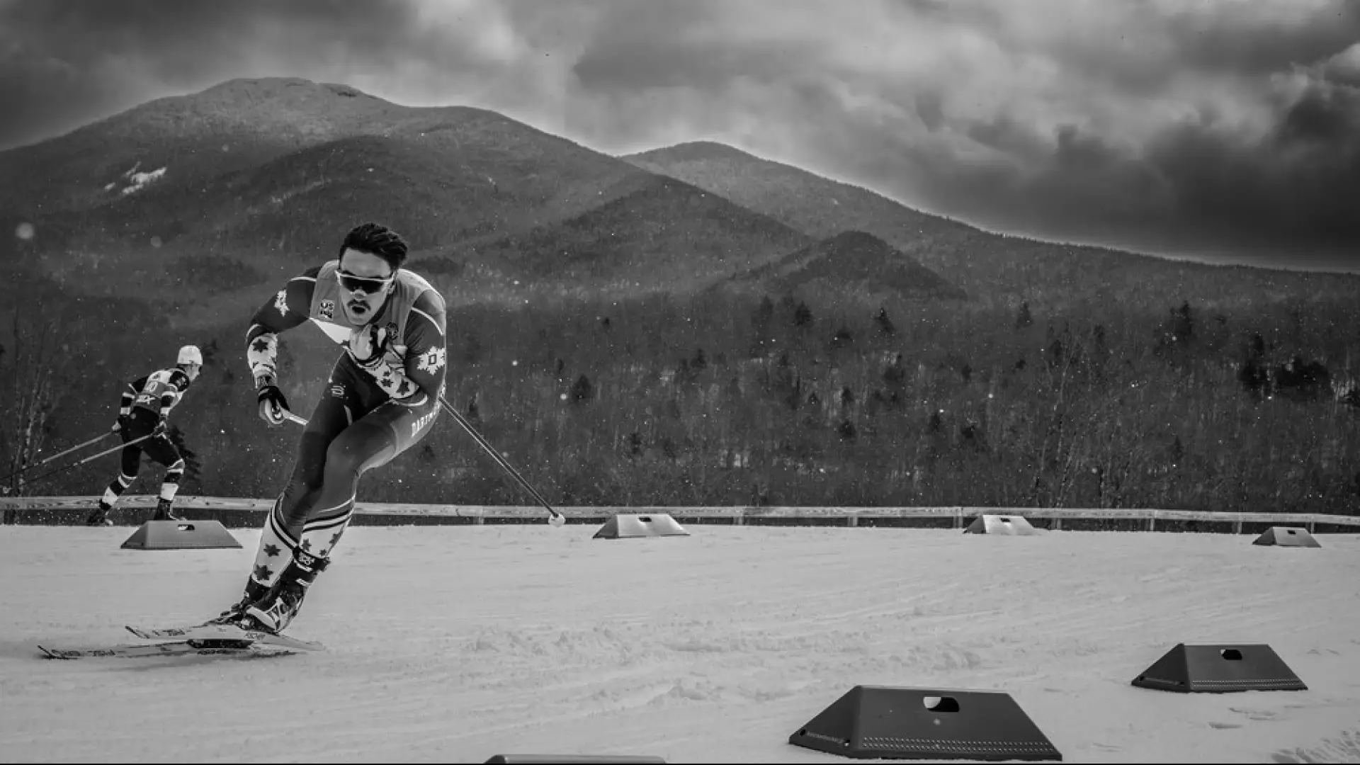 Black and white image of two men cross-country skiing on a competition trail with mountains in the background