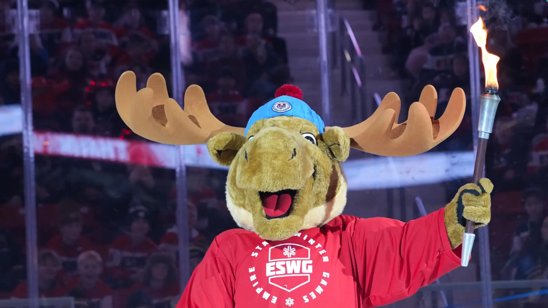 A moose mascot wearing a red Empire State Winter Games jersey holds a flaming torch aloft during an opening ceremony.
