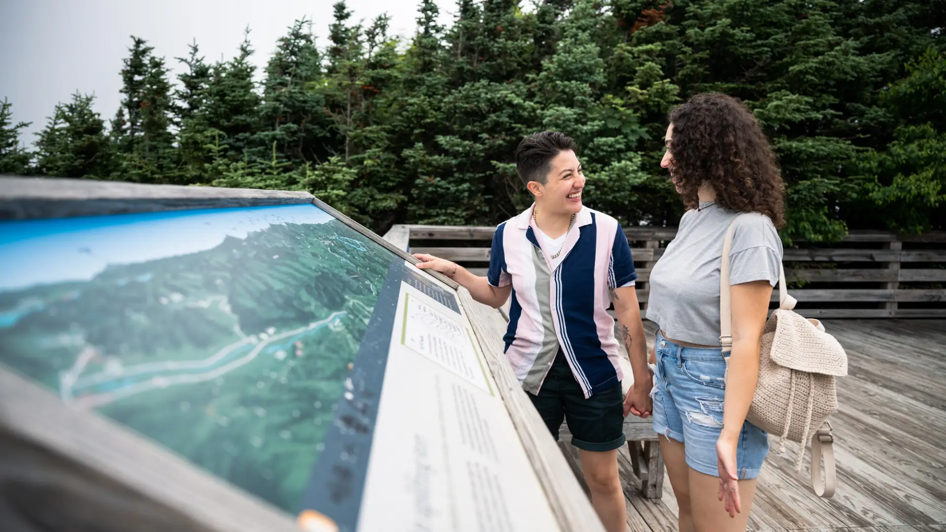 A couple stands smiling at each other in front of an interpretive display on a deck with forest in the background.
