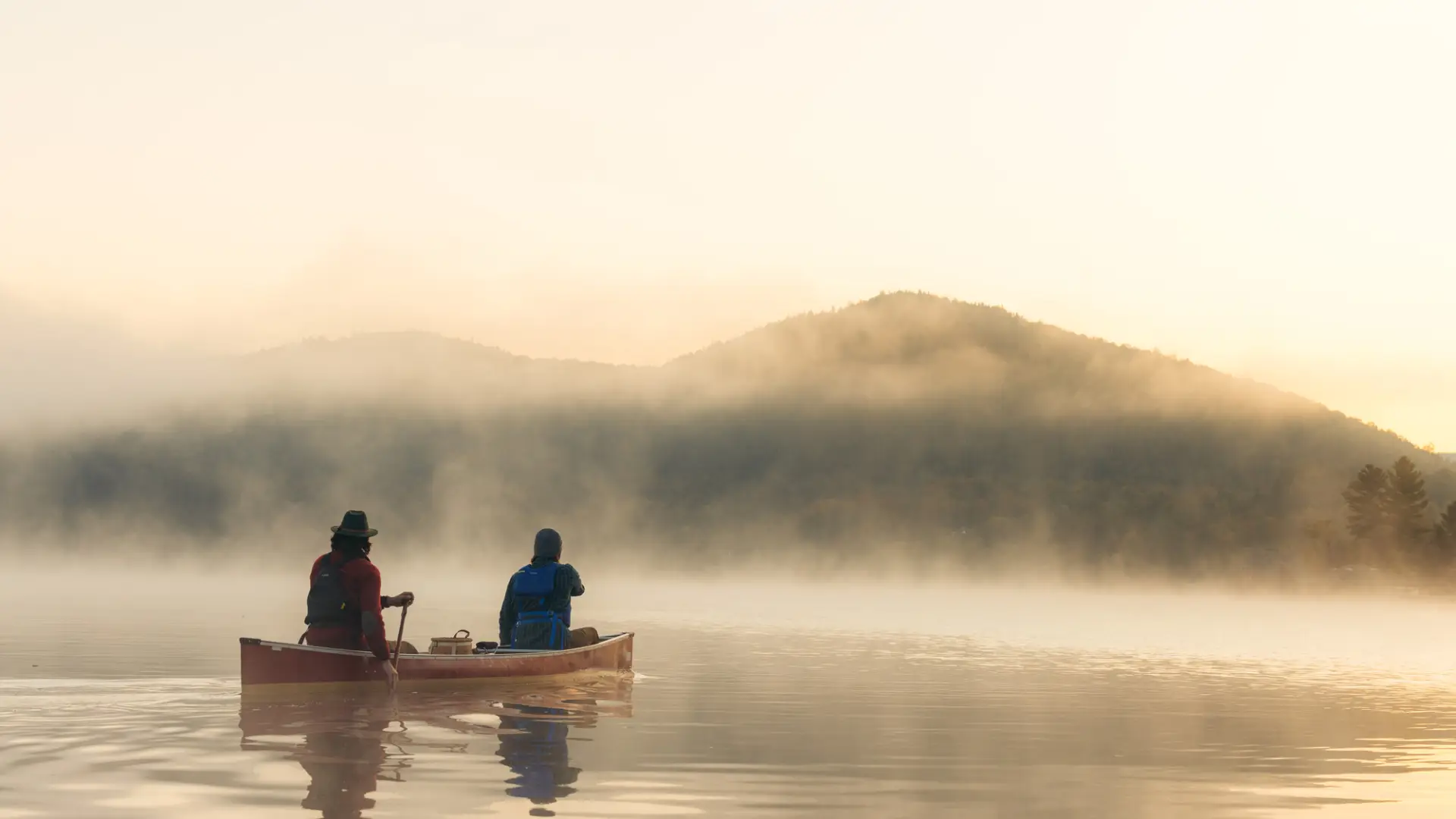 Two people, one wearing a wide brimmed hat, paddle away from the camera on a lake toward mountainous shoreline on a misty morning