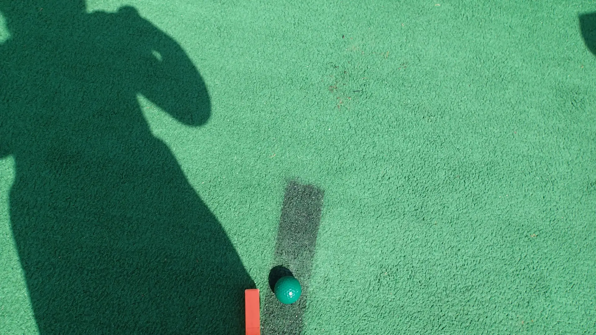 An image of artificial turf on a mini golf course, with a putter, golf ball, and the shadow of a little girl.