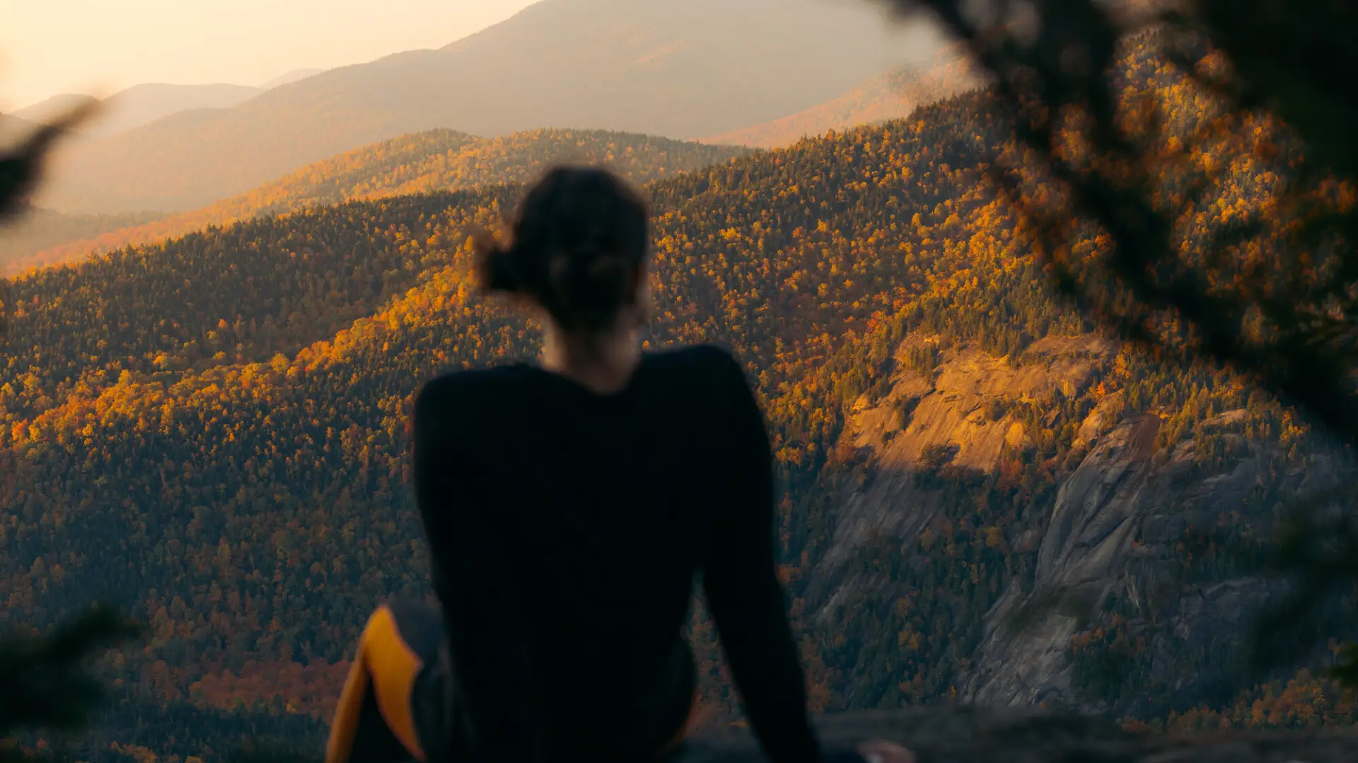 A person sitting with their back to the camera, looking out over a vast, layered landscape of mountains and forests colored in the gold and orange hues of autumn at sunset.