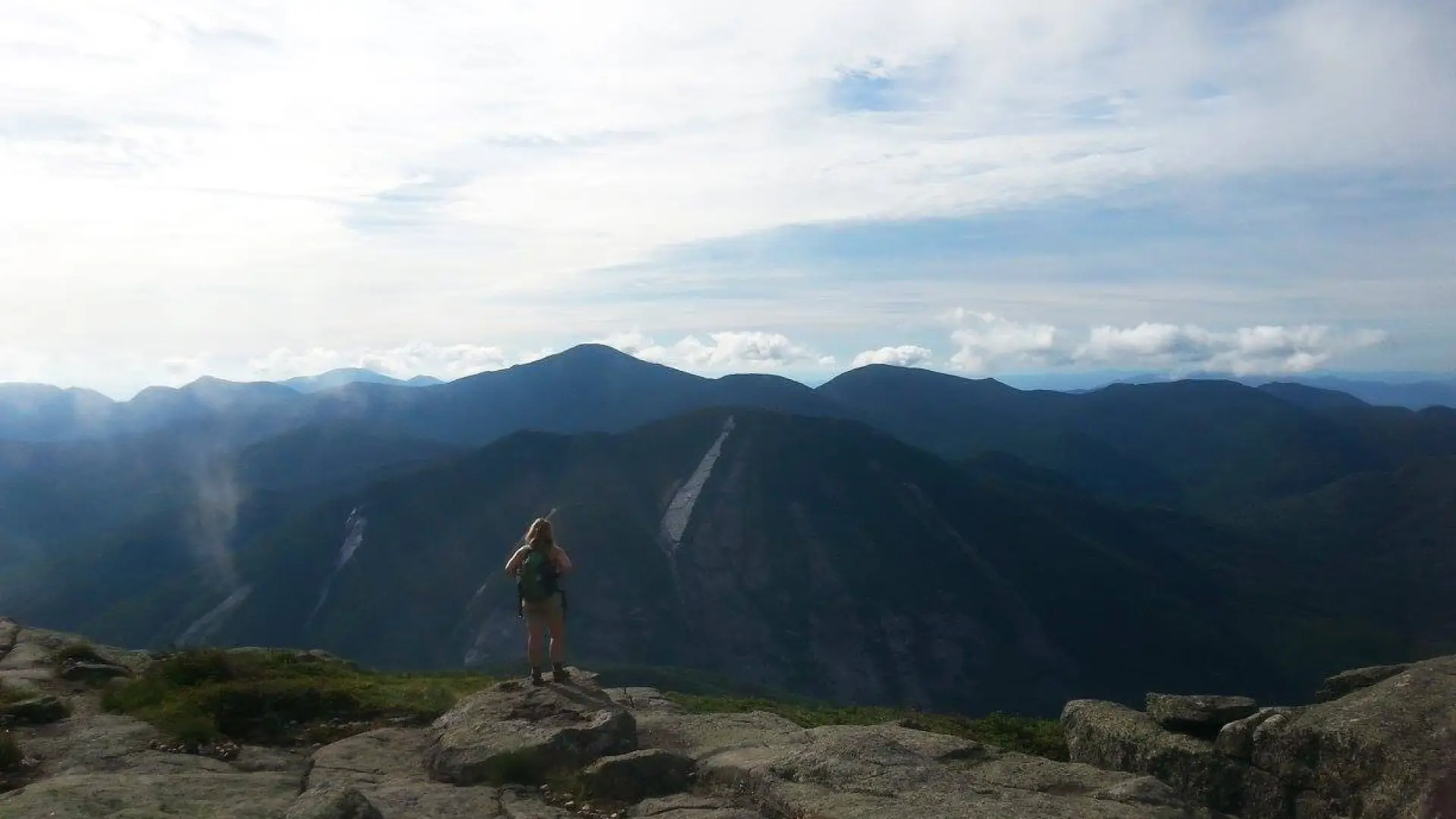 A woman with hiking backpack stands admiring the view in the High Peaks.