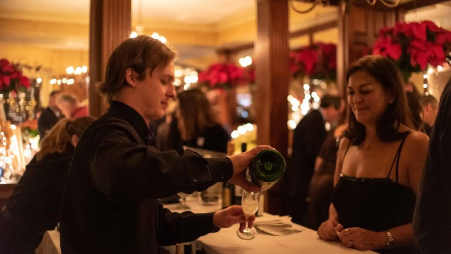 Bartender in dress clothes pours sparkling wine into a flute glass for a woman in a black dress on the other side of the bar at a busy party