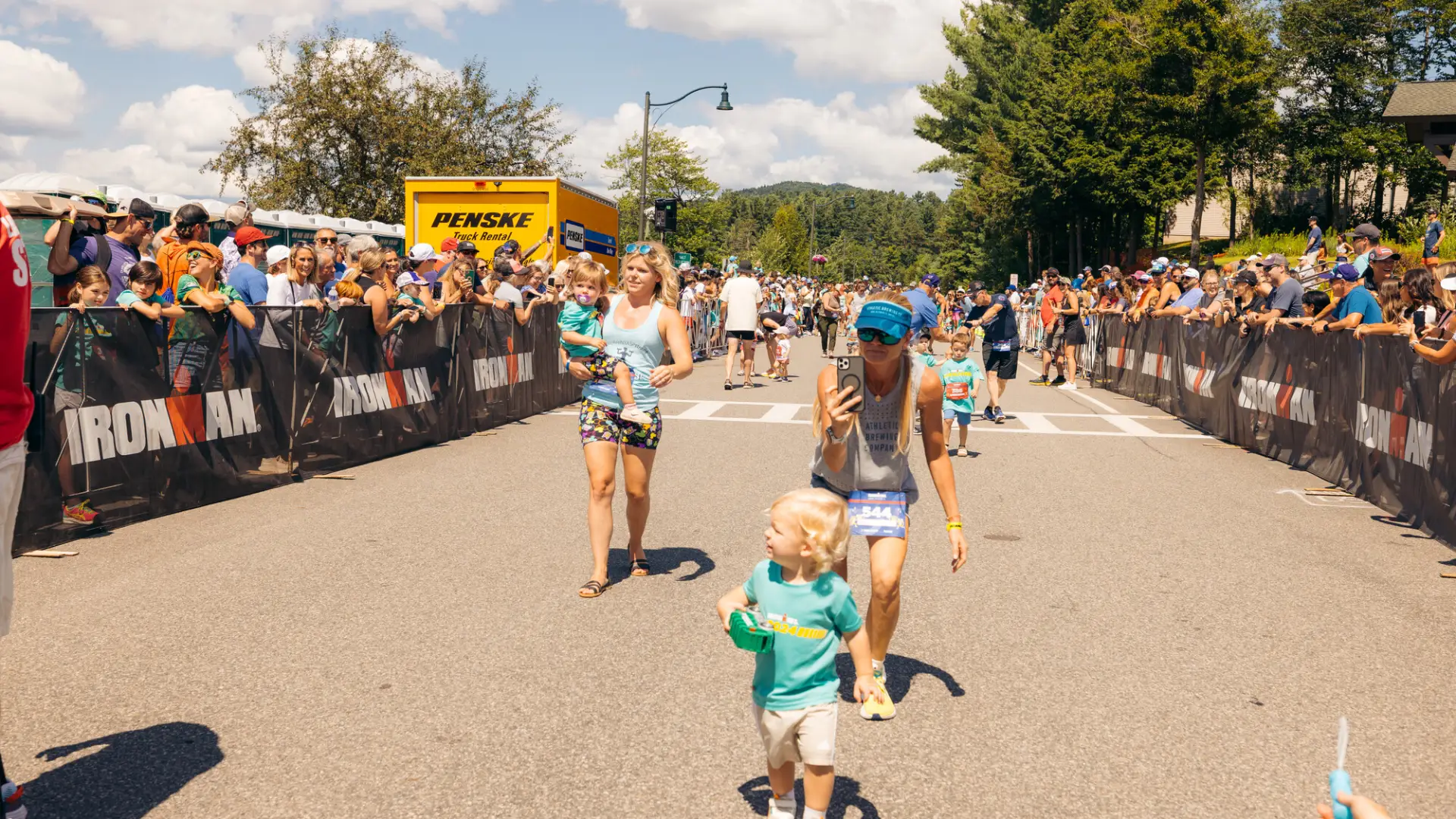 Woman walks behind child filming them and second woman carries toddler on Ironkids race course while spectators cheer