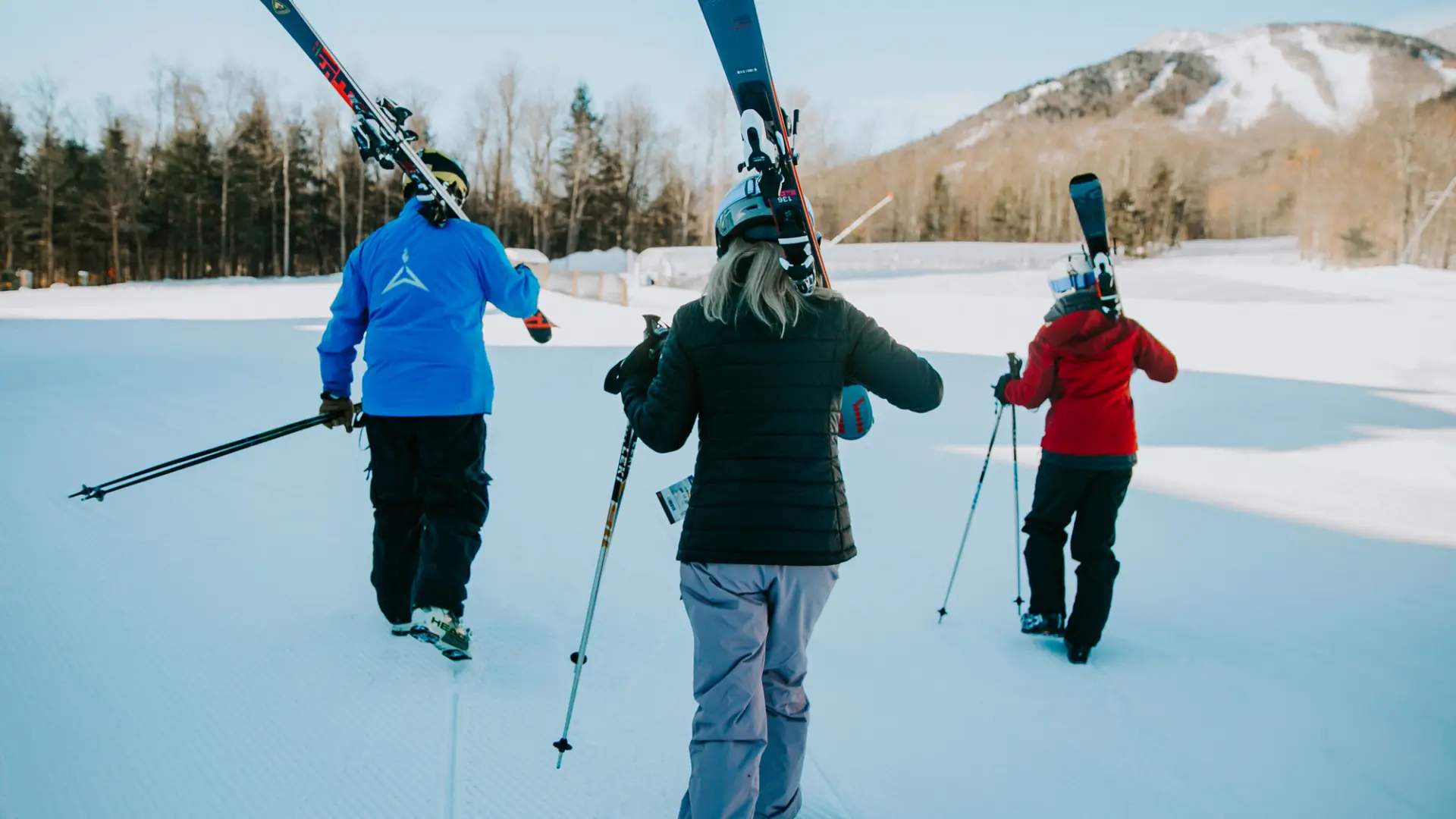 Three skiers in blue, black, and red jackets walk across a groomed trail carrying their skis over their shoulders.