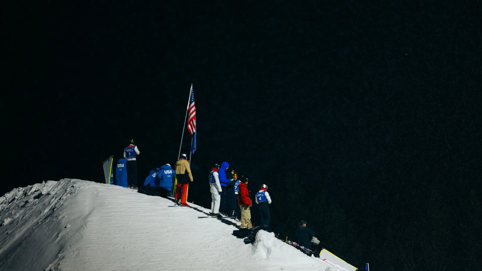 Group of people and skiers stand atop a snowy hill at night, illuminated with artificial light, looking outward into the dark empty space in front of them