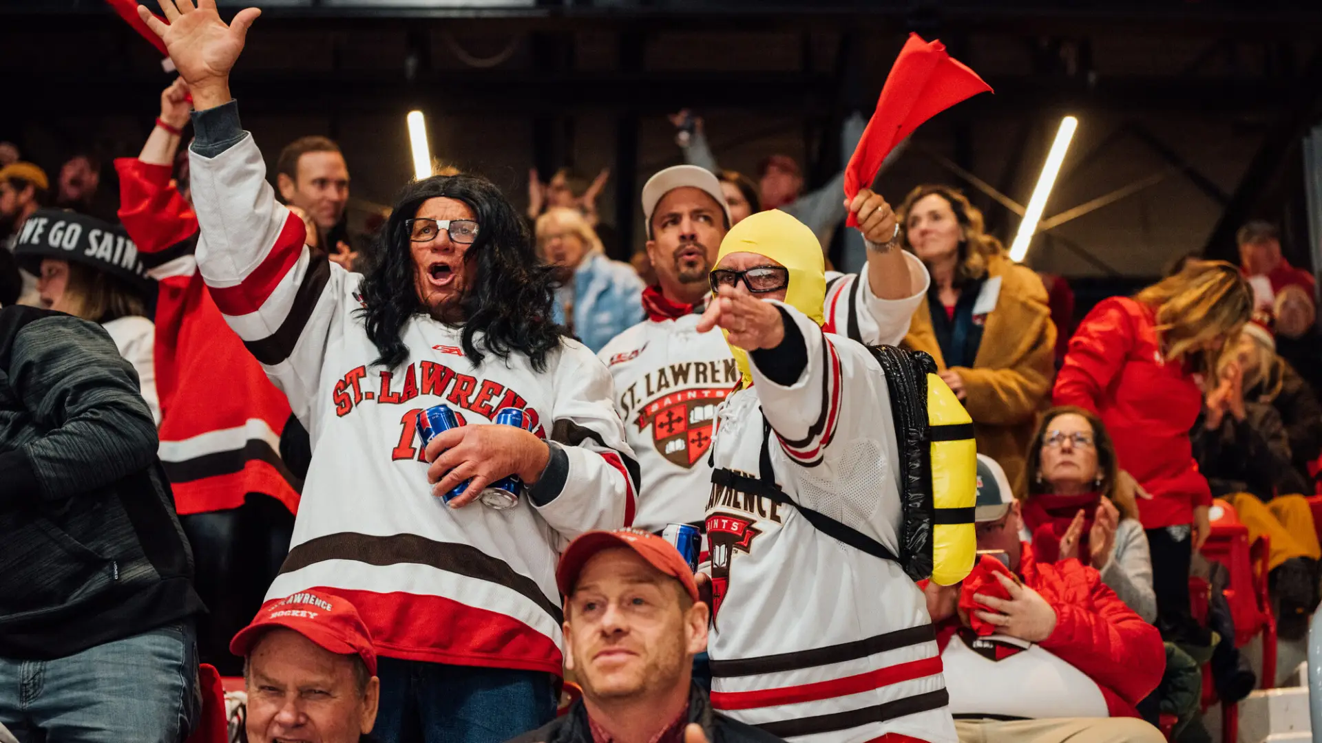 St Lawrence University hockey fans are seen standing in seats during game wearing jerseys and other costume items, cheering enthusiastically