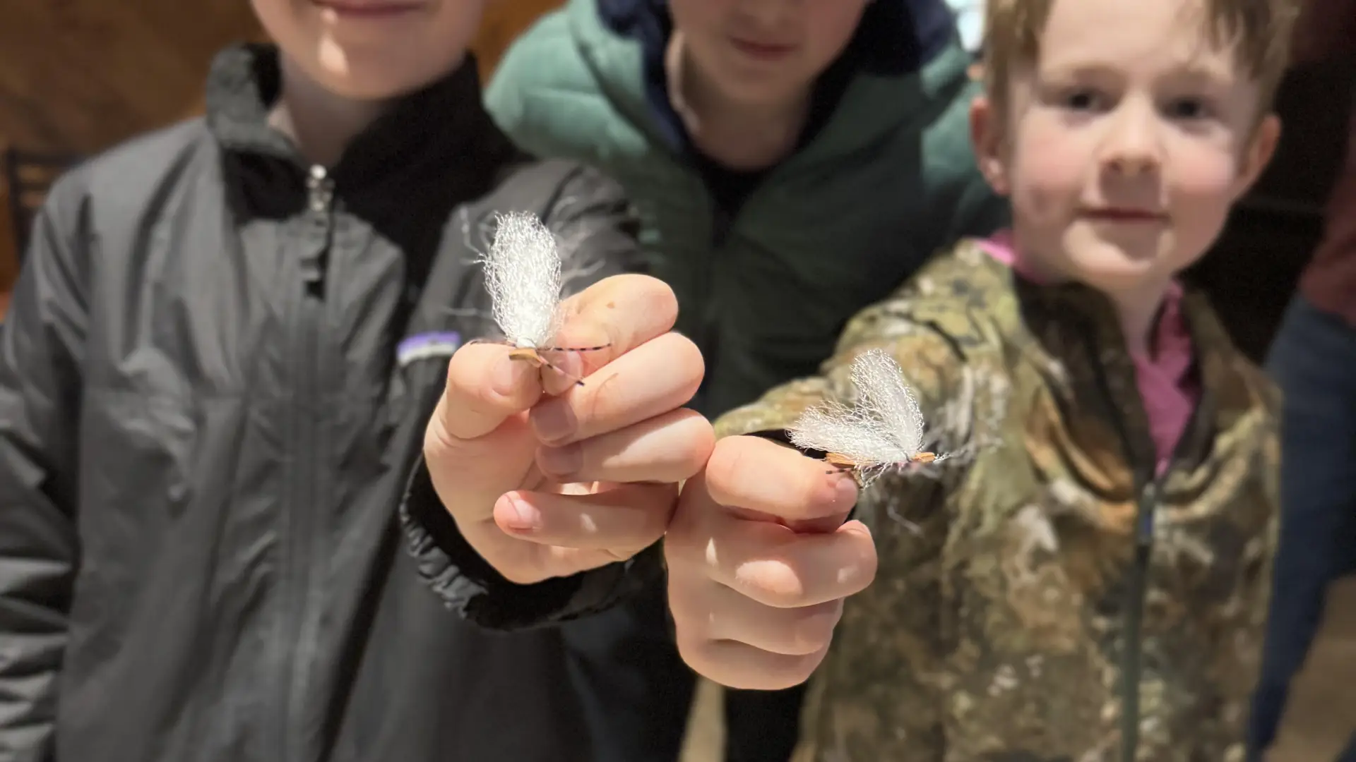 Two kids holding fly fishing flies up to camera while third child looks over their shoulders