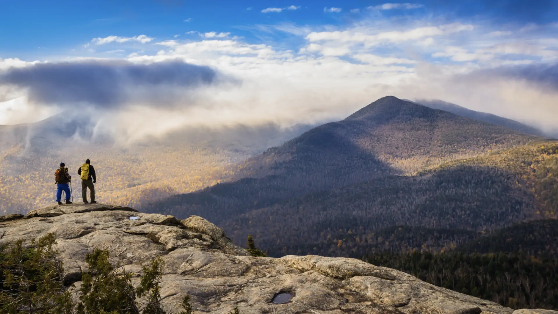 Two hikers stand atop a rocky mountain summit in the Adirondacks in fall looking out at view of other mountains